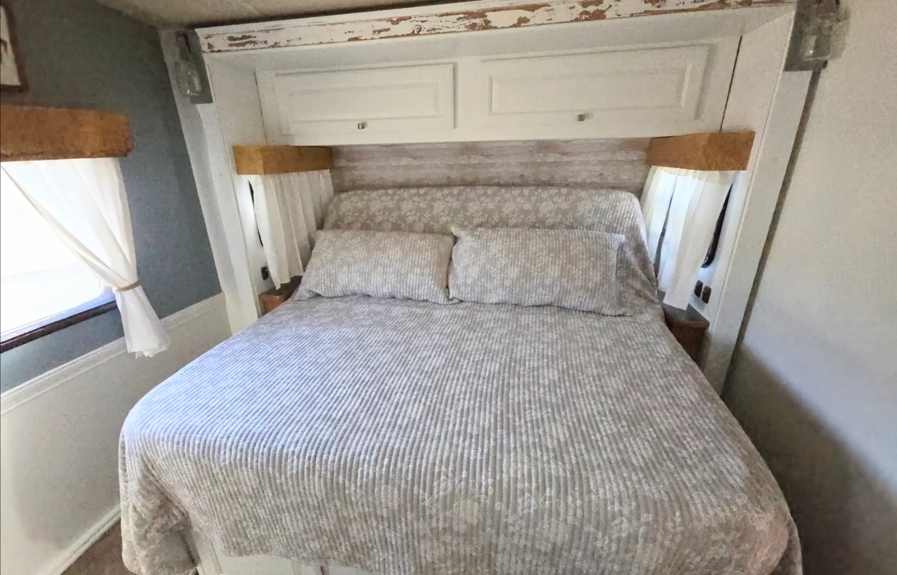 Cozy RV rental bedroom featuring floral bedding, white overhead cabinets, and a window with tie-back curtains. Perfect for travel.. Itasca Sunova 2014