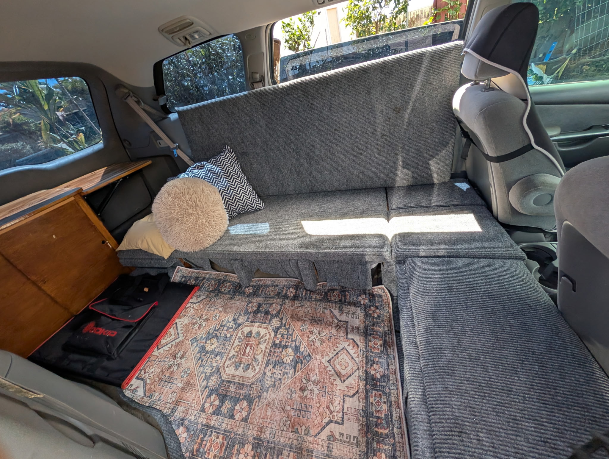 Cozy RV rental interior: custom van conversion with a gray convertible bed, pillows, wooden storage, and a patterned floor rug.. Toyota Trailfinder 2008