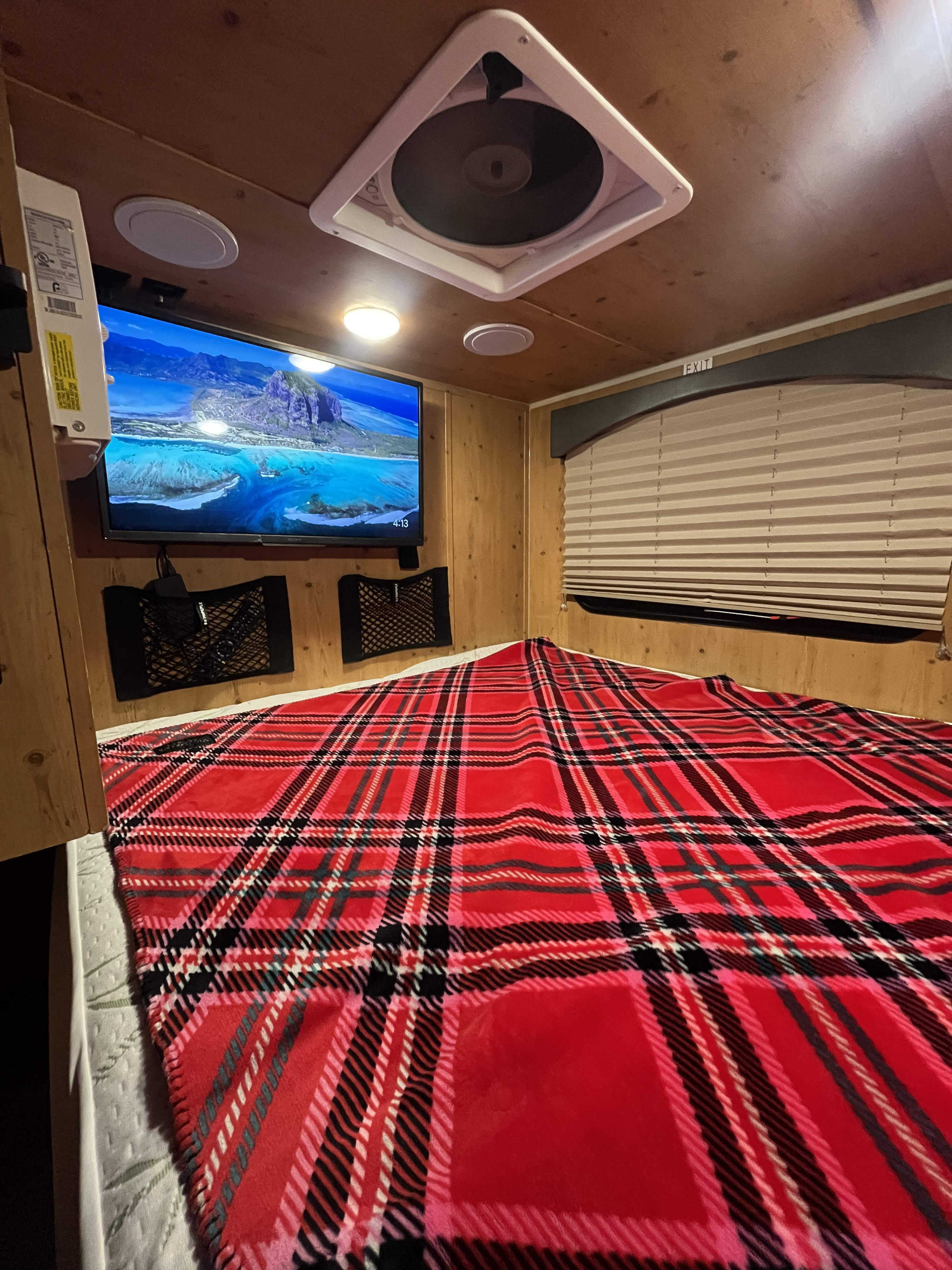 Cozy RV rental bedroom featuring a bed with a vibrant red plaid blanket, a mounted TV, wood paneling, and a window with blinds.. Sunset Park RV SunRay 2021