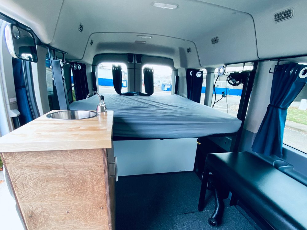 Spacious RV rental camper van interior with a comfy bed, functional kitchenette featuring a sink, and a cushioned bench seat.. Ford Transit 2019