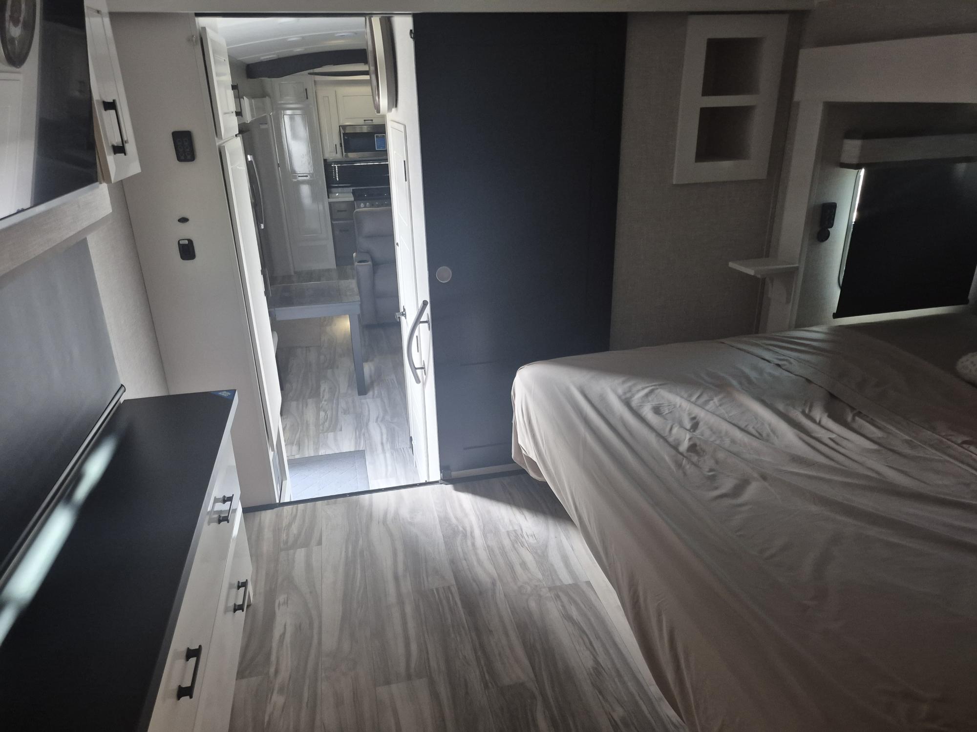 Inside a modern RV rental: a bedroom with a cozy bed, dresser, and a sliding door revealing the kitchen/living space.. Jayco Pinnacle 2025