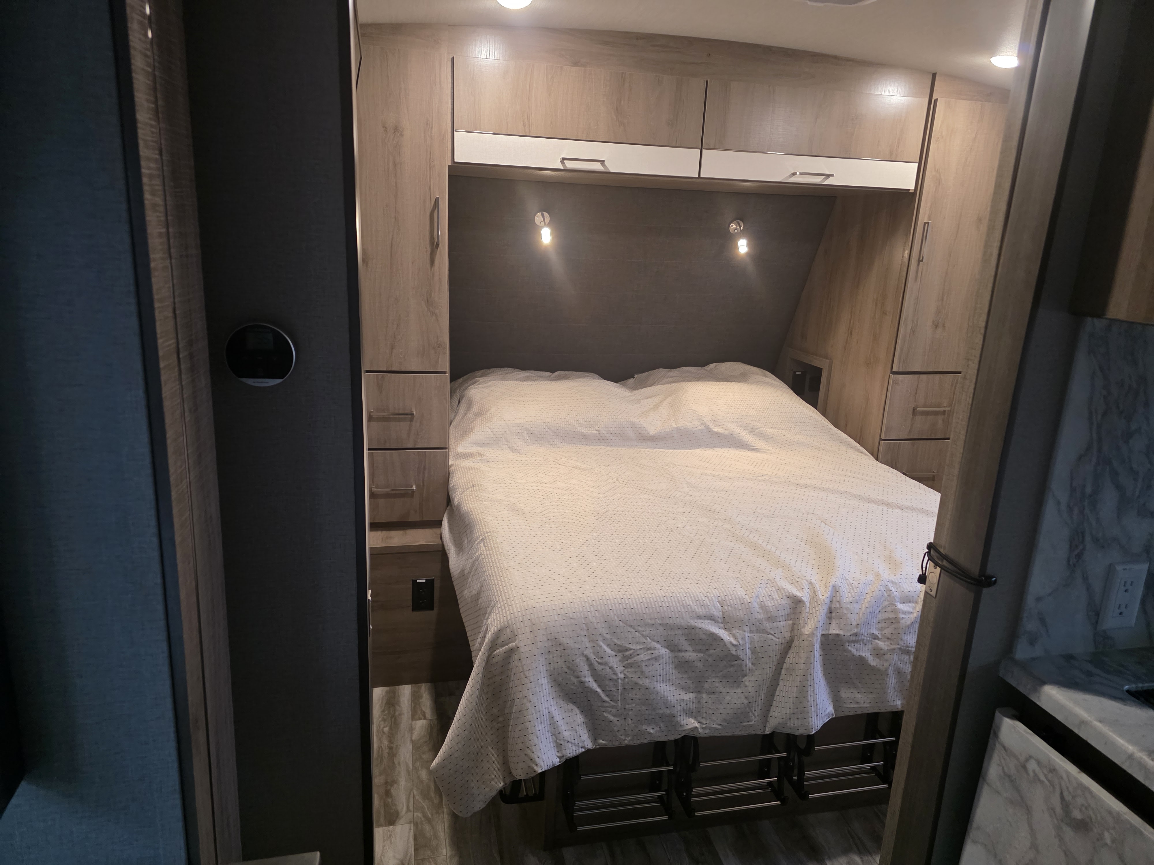 An RV rental's cozy bedroom area, featuring a bed, wood-look cabinets, and sconce lighting.. Grand Design Imagine 2022