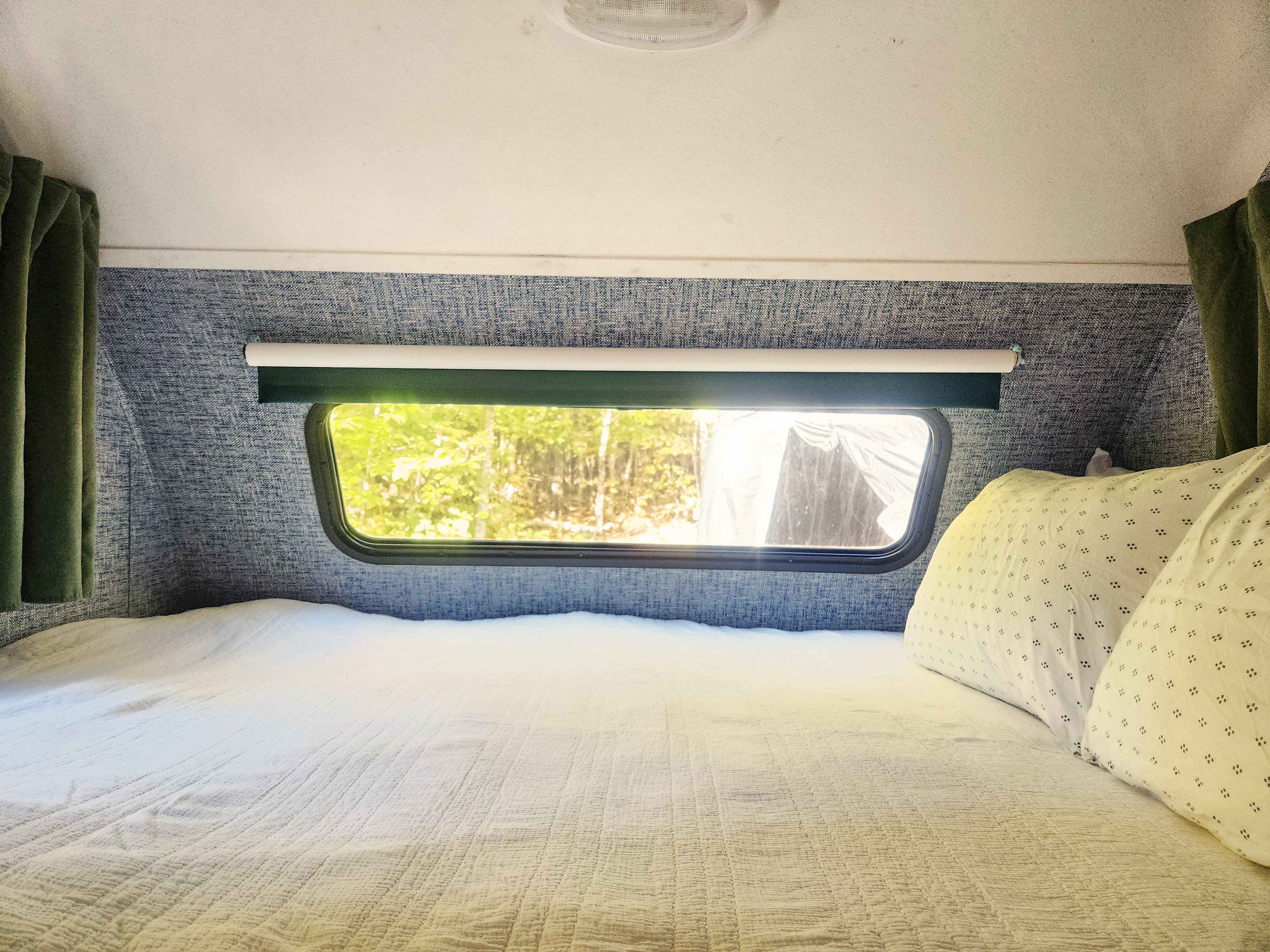 Inside a cozy RV rental: a comfortable bed with white covers and patterned pillows, with a window framing a tranquil forest view.. Forest River R-Pod 2010