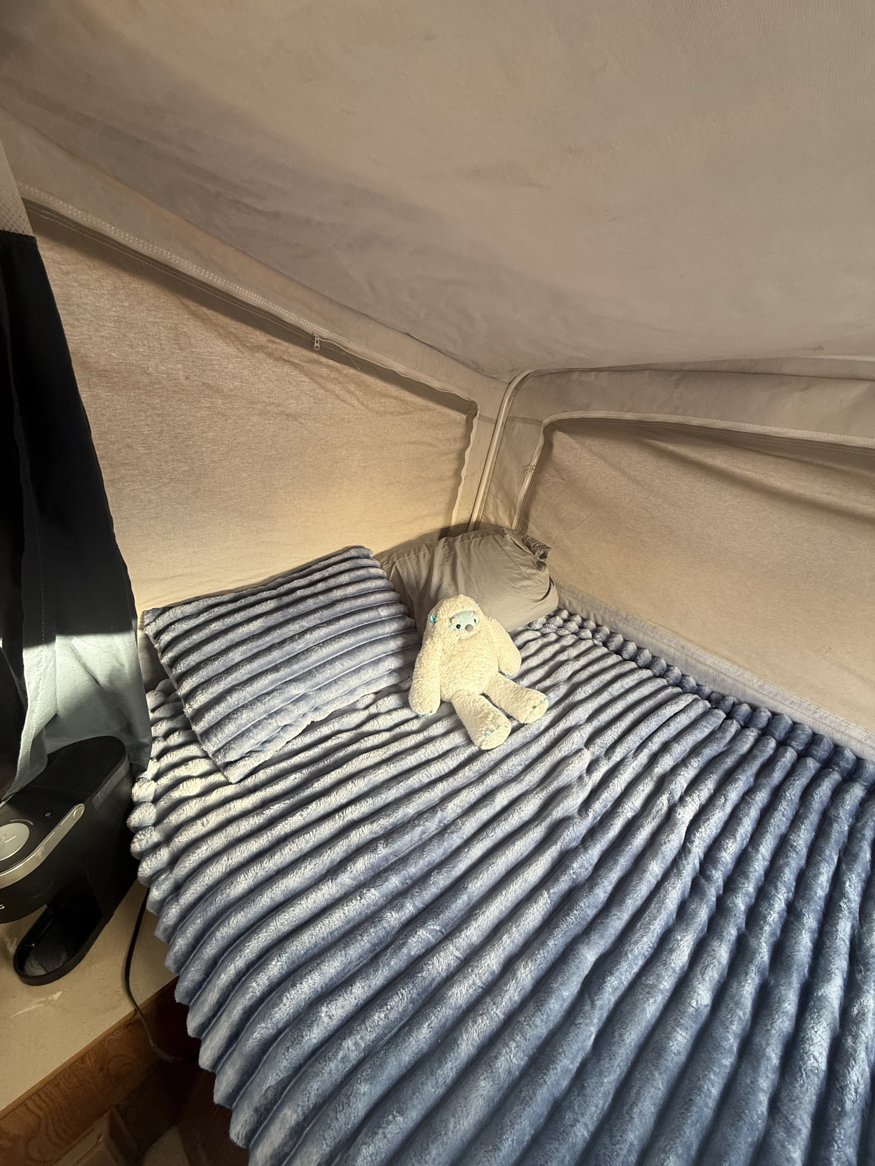 A cozy RV rental sleeping area with a blue ribbed blanket, pillow, and a fluffy white toy, set within a pop-top camper.. Dutchmen Voyager 2000