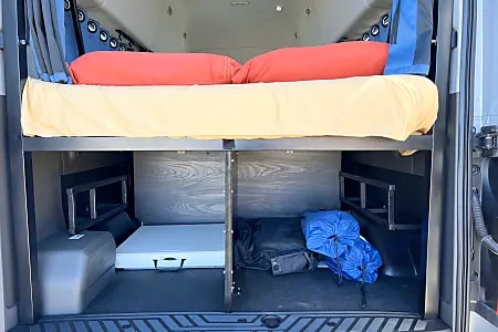 RV rental interior: A raised bed with orange pillows above organized under-bed storage for gear and a folding table.. Ford Transit 2019