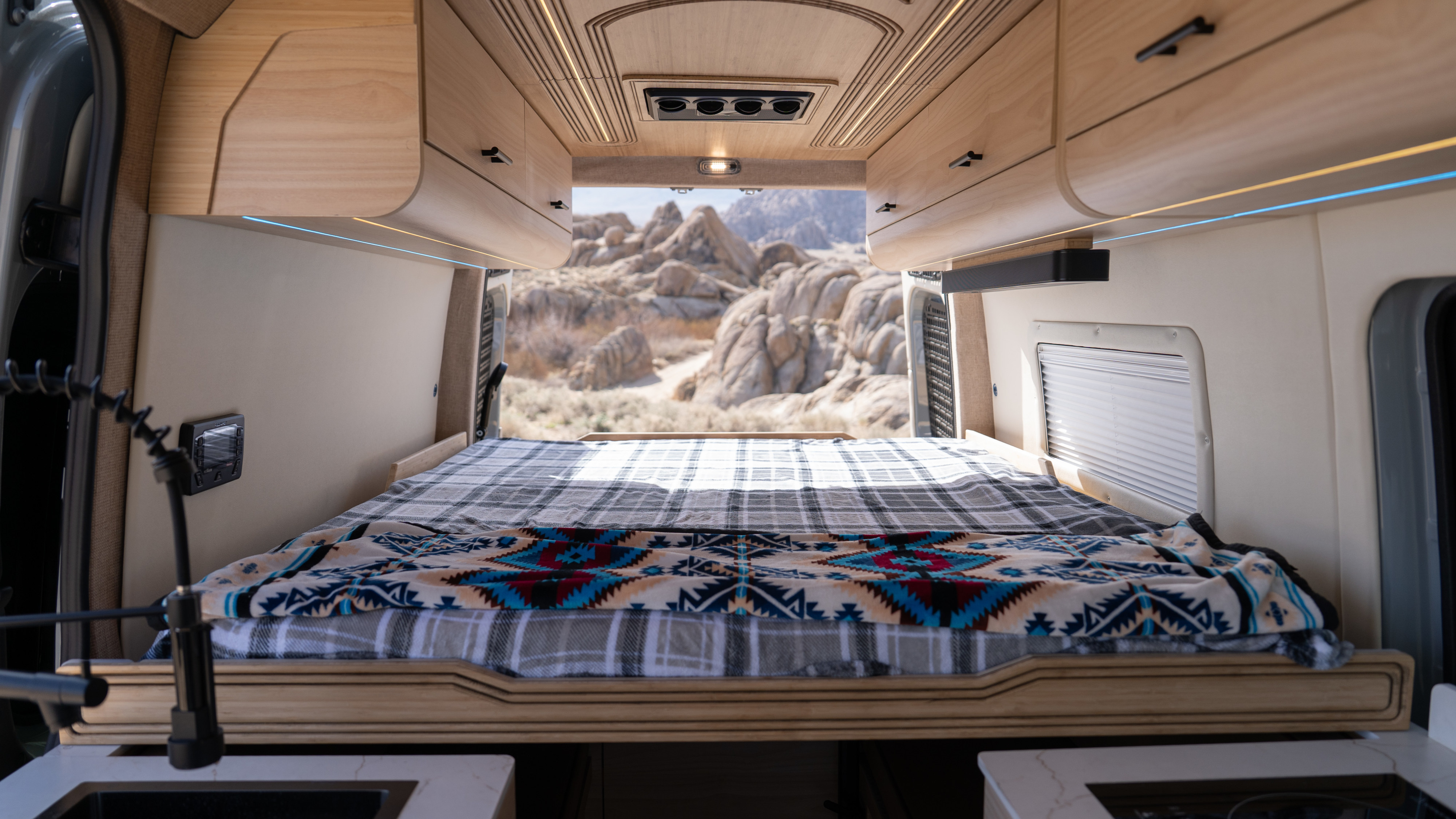 Inside an rv rental, a bed with colorful blankets offers a desert mountain view from the open rear doors, showing the comfortable living space.. Other Mercedes Sprinter 2024