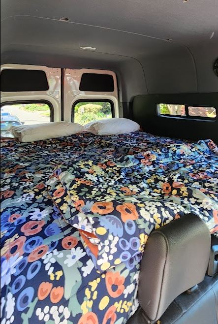 Cozy RV rental camper bed with a vibrant floral comforter and pillows, viewed from inside the van with windows.. Mercedes Sprinter 2023