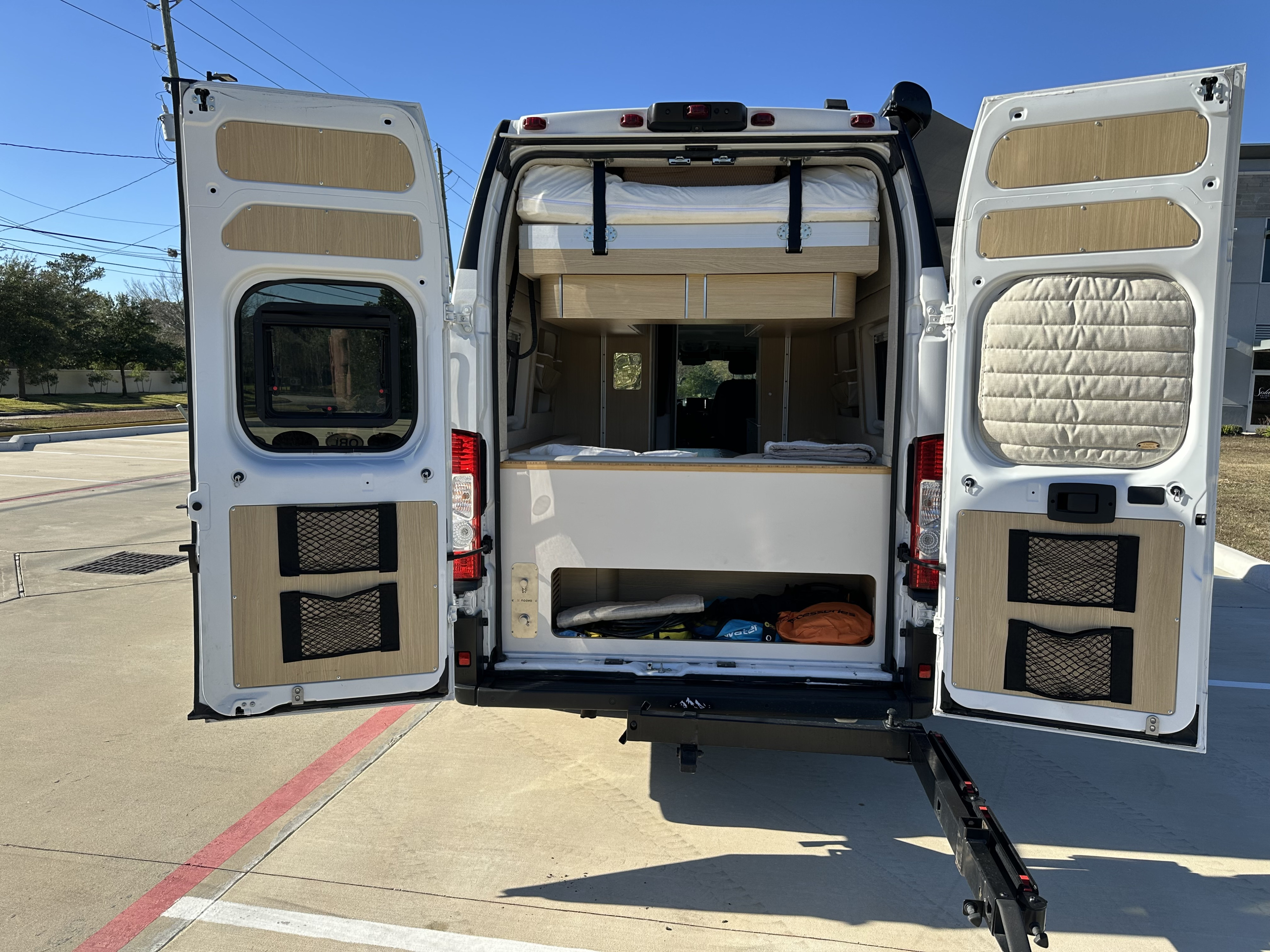 Rear view of an RV rental camper van with open doors, revealing a custom interior with an upper bed, lower storage, and modern wooden details.. Noovo Noovo Class B 2024
