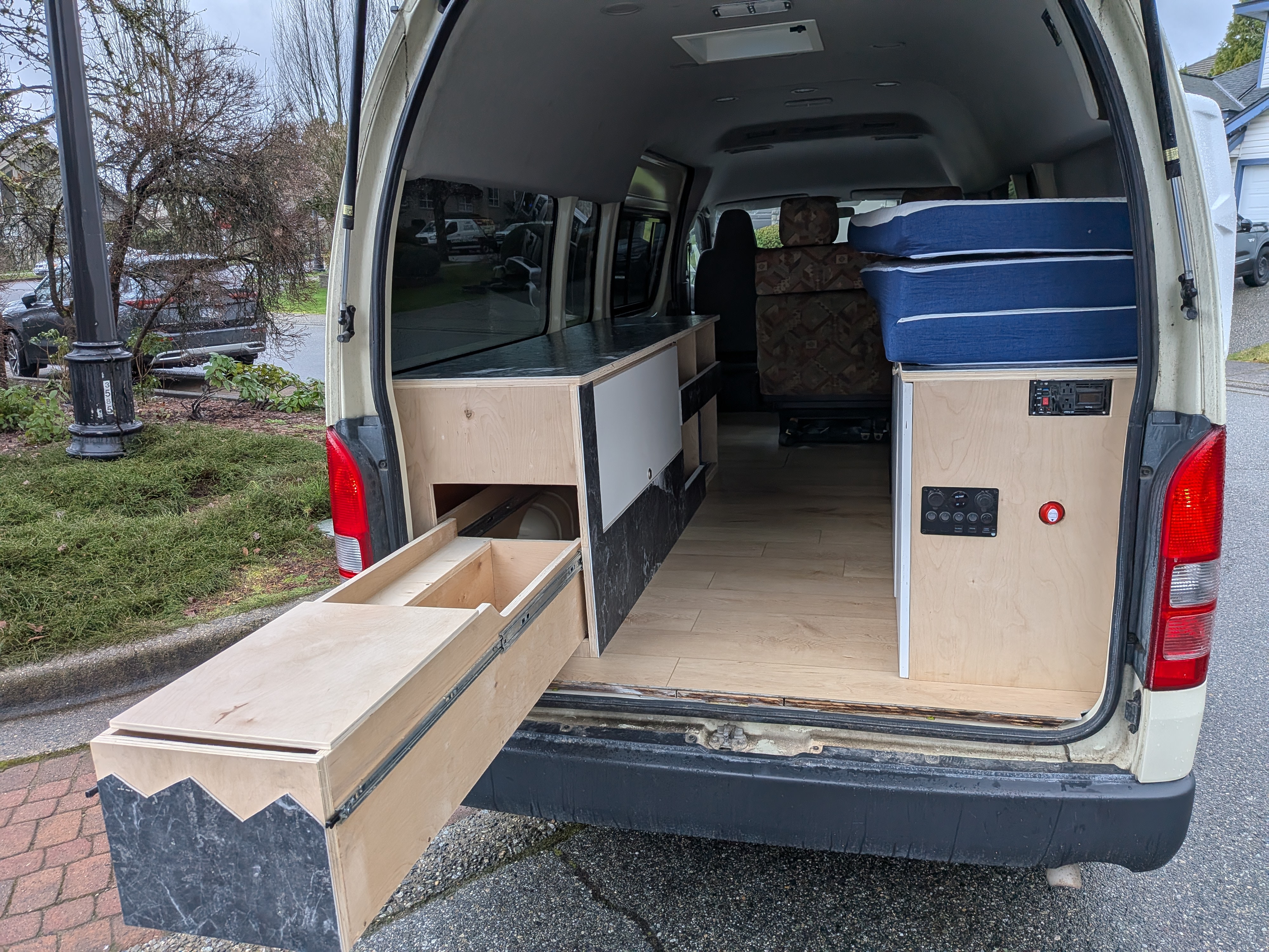 Rear view of a DIY camper van interior, ready for rv rental, with custom cabinetry, a pull-out bed & control panel.. Toyota Hiace 2006