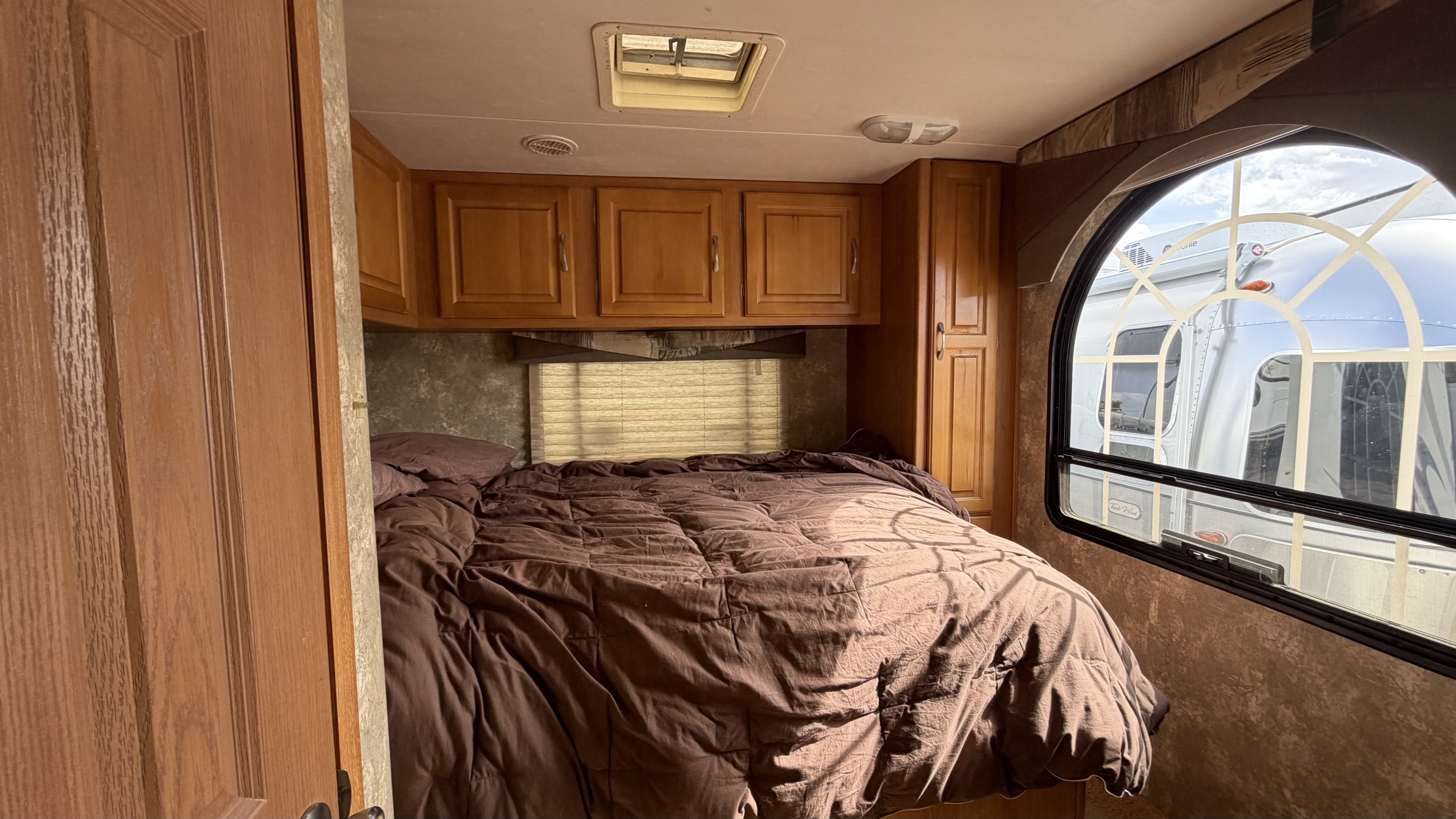 Cozy RV rental bedroom with a large brown bed, wooden cabinets, skylight, and a window showing another RV outside.. Coachmen Freelander 2007