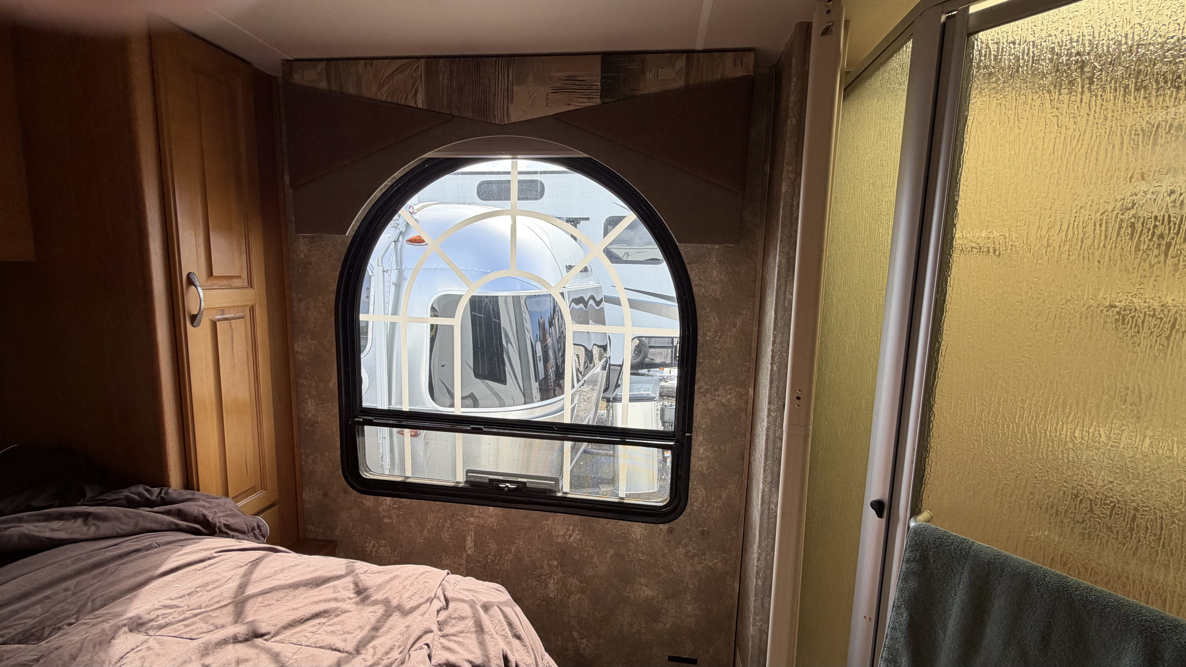 Inside an RV rental bedroom: brown bed, wooden cabinets, arched window looking out at another RV, and a textured shower door.. Coachmen Freelander 2007