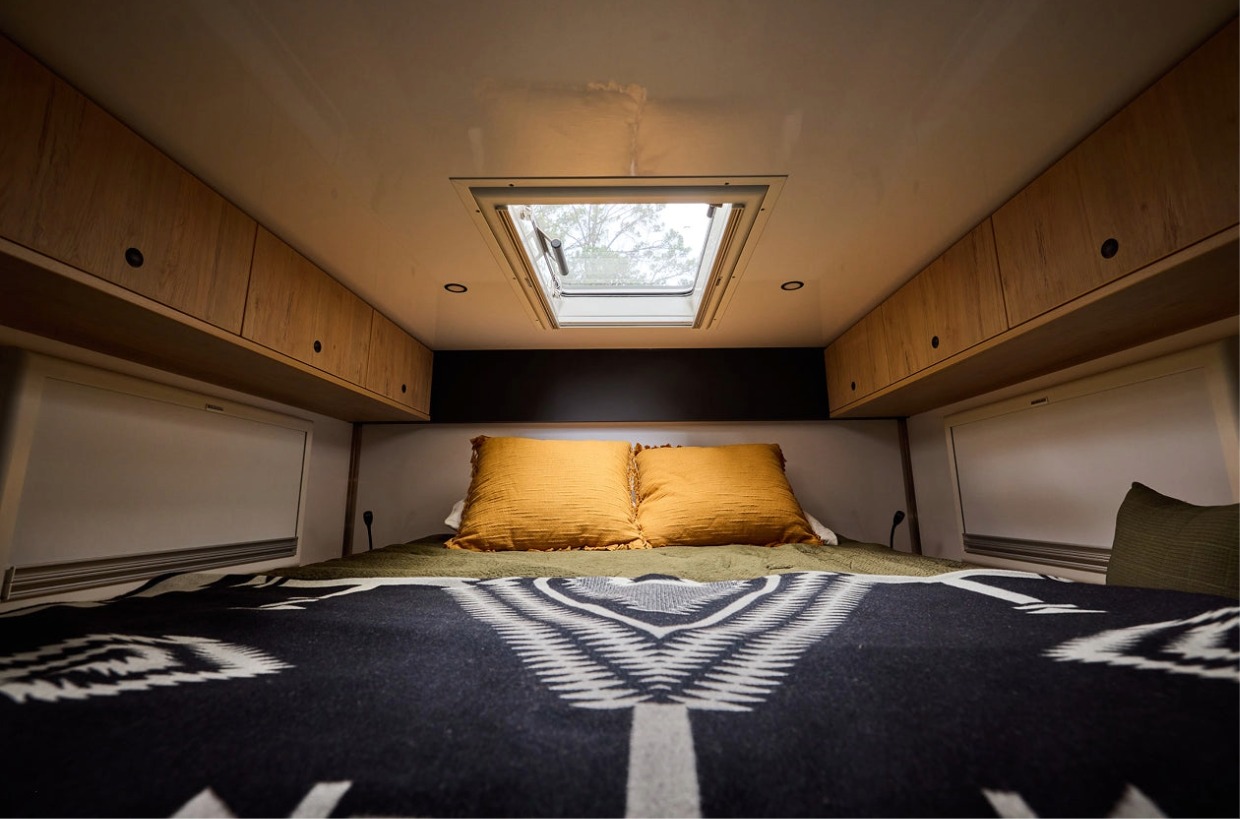 Cozy RV rental bedroom: bed with yellow pillows, patterned blanket, skylight, and wood overhead cabinets.. Storyteller Epic 2025