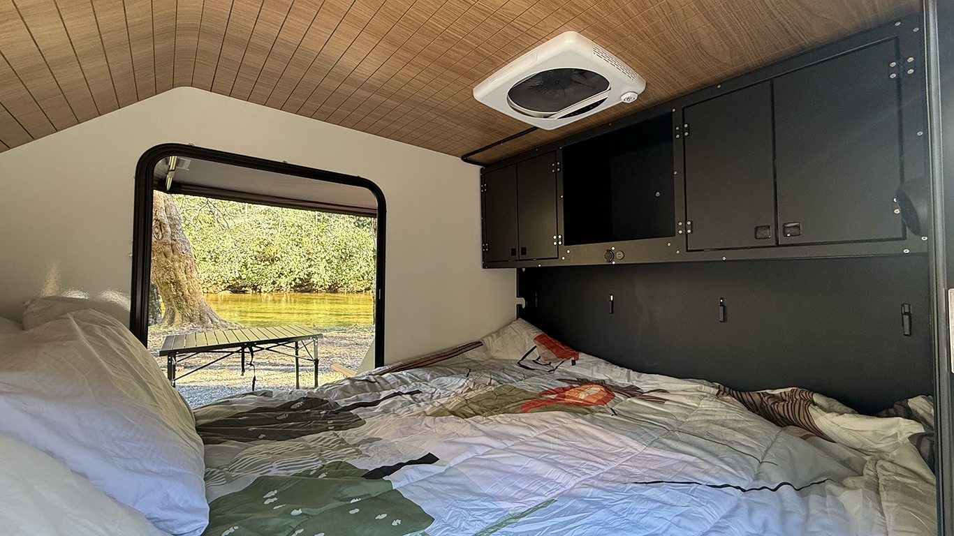 Cozy RV rental interior with bed, overhead storage, and a scenic view of a river/lake and trees outside the open door.. Cedar Ridge Deneb XT 2025