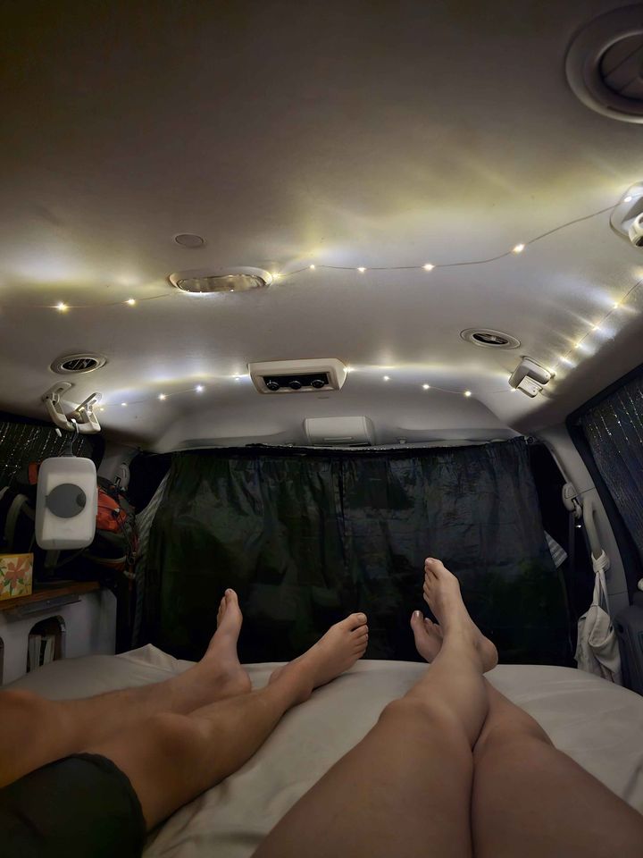 Inside a cozy RV rental, two people relax with their feet up on the bed under soft string lights, perfect for van life adventures.. Dodge Grand Caravan 2009