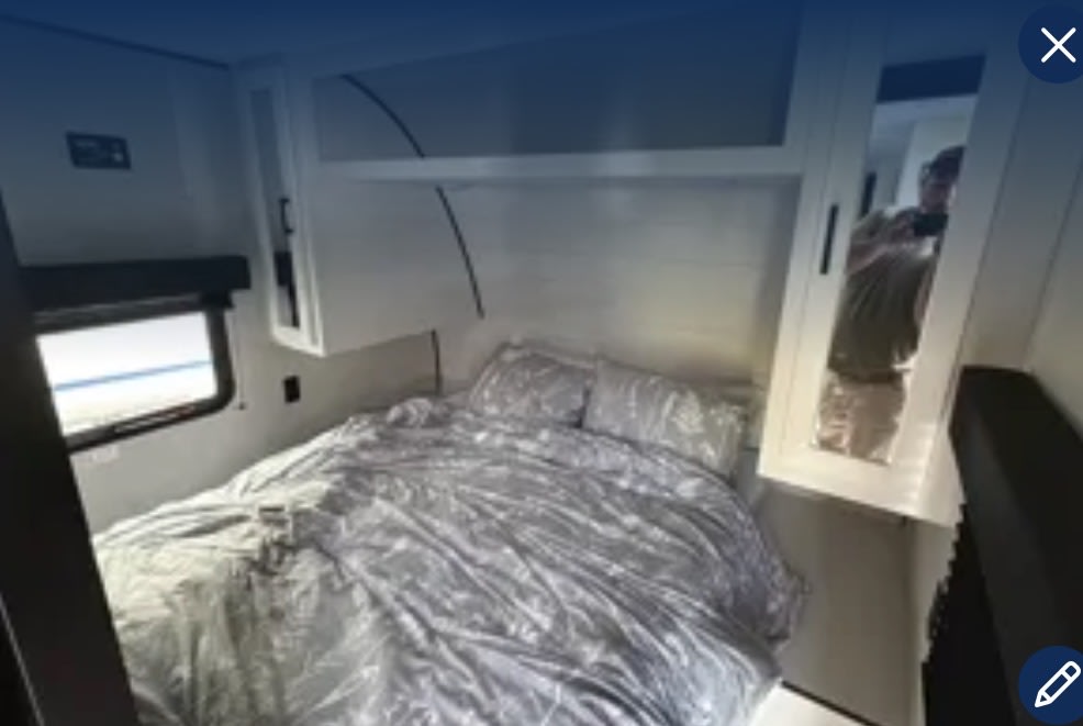 Interior of an RV rental bedroom showing a bed with gray bedding, overhead cabinets, and a window. Reflecting person in mirror.. Jayco Jay Flight SLX 2024
