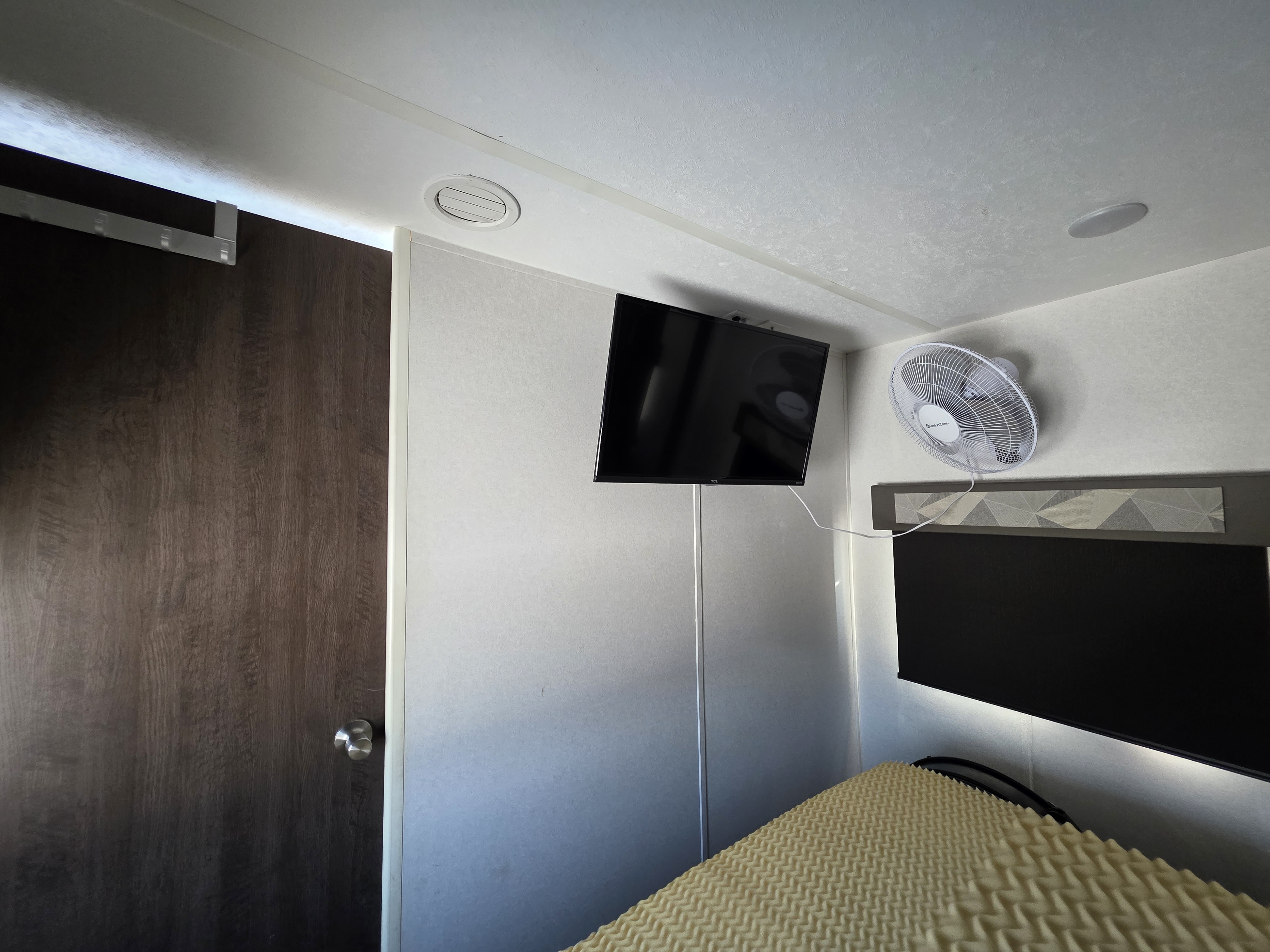 Cozy RV rental bedroom featuring a wall-mounted TV & fan, and a textured foam bed. A dark door is on the left.. Forest River Salem 2019