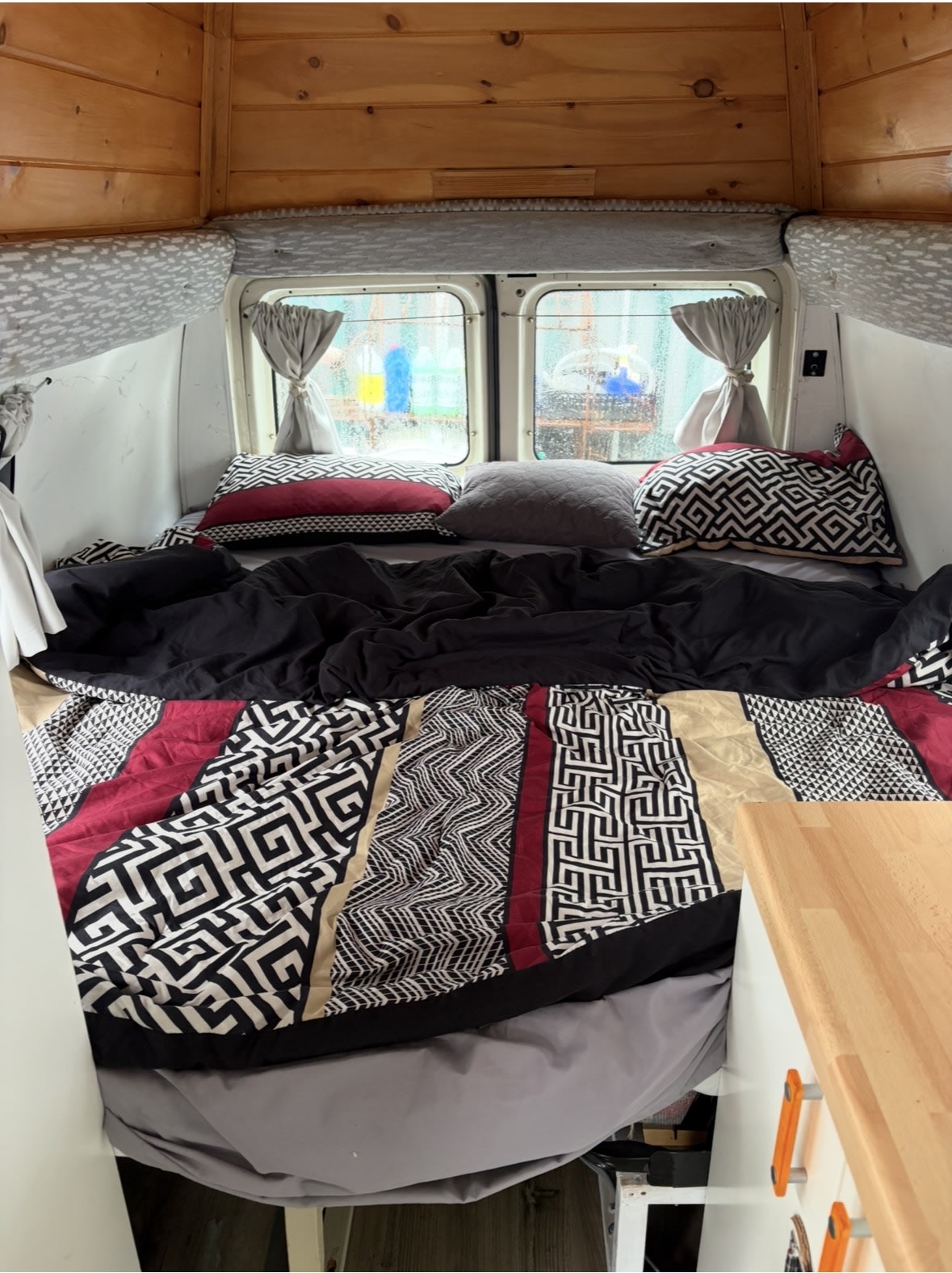 Cozy rv rental interior with patterned bed, pillows, and wooden accents, seen through rainy windows.. Ford Econoline Club Wagon 1994