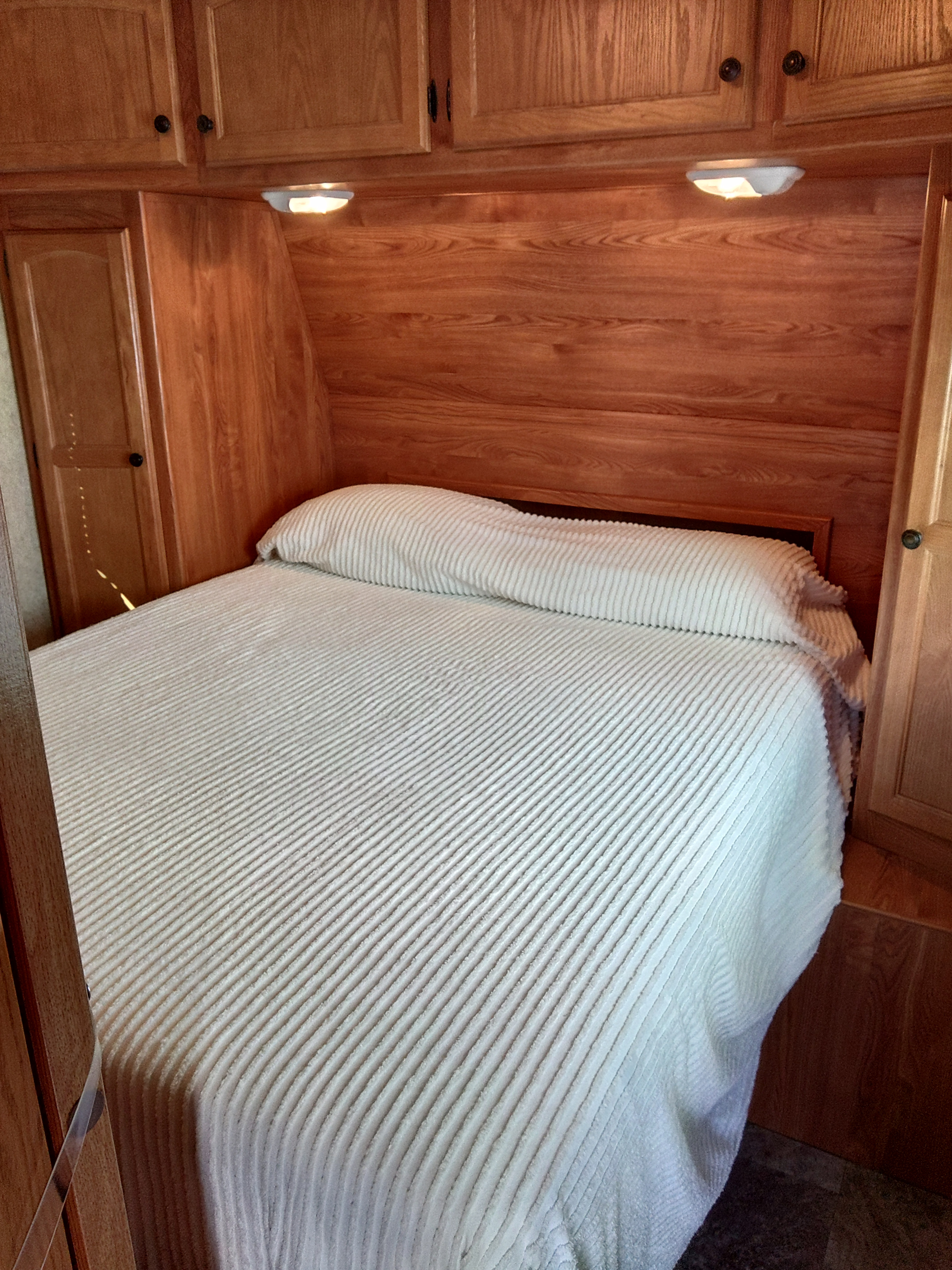 Cozy RV rental bedroom featuring a striped white bed, wood paneling, overhead lights, and cabinets for a comfortable stay.. Heartland RVs Trail Runner 2012