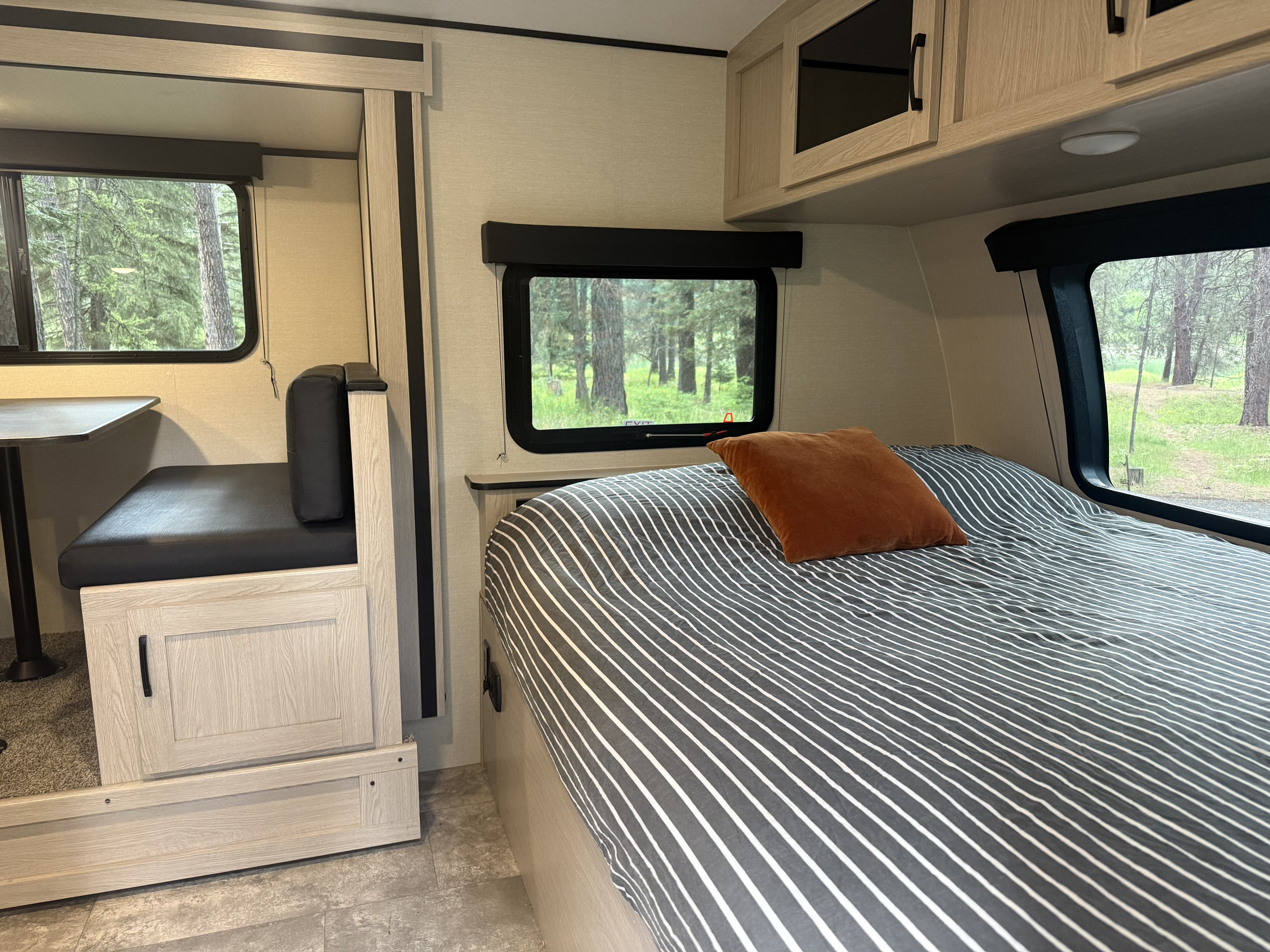 RV rental interior: Cozy bedroom with striped bedding & dinette, windows reveal a serene forest view.. Coachmen Apex Nano 2023
