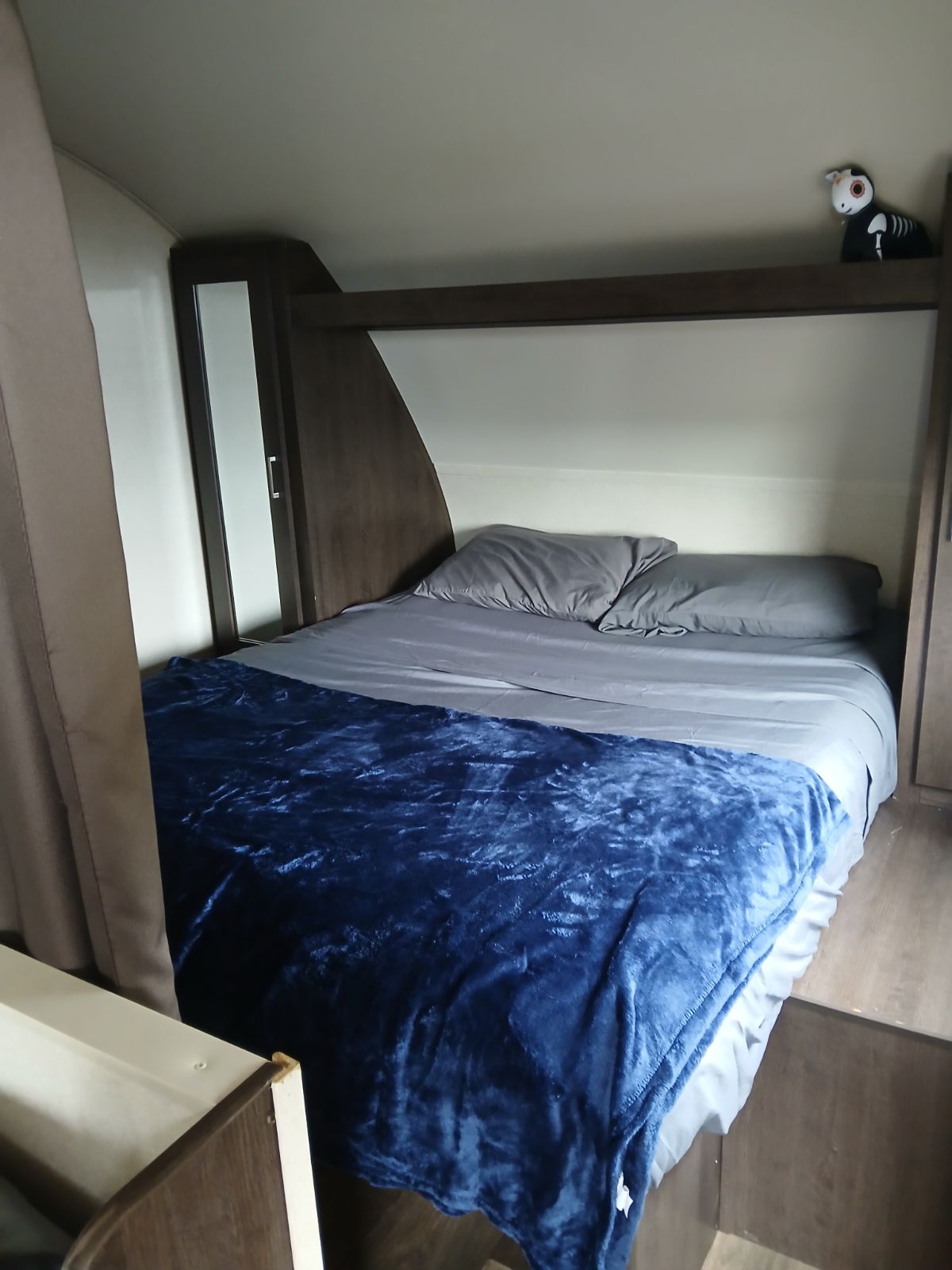 Inside an RV rental: a compact sleeping area featuring a bed with gray sheets, a plush blue blanket, and a fun skeleton toy on a shelf.. Xlite 261BHXL 2019
