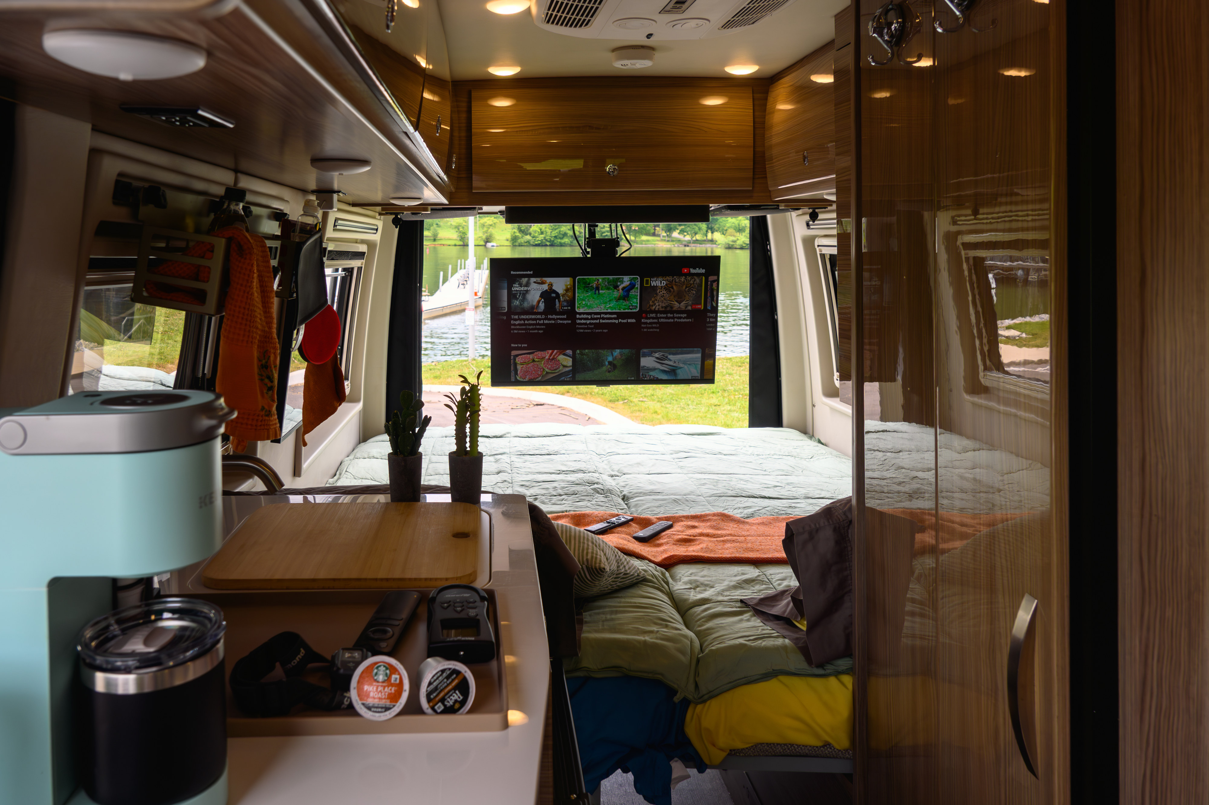 This RV rental interior features a cozy bed with a scenic lake view, TV playing YouTube, coffee maker, and organized kitchen essentials.. Winnebago Boldt 2022