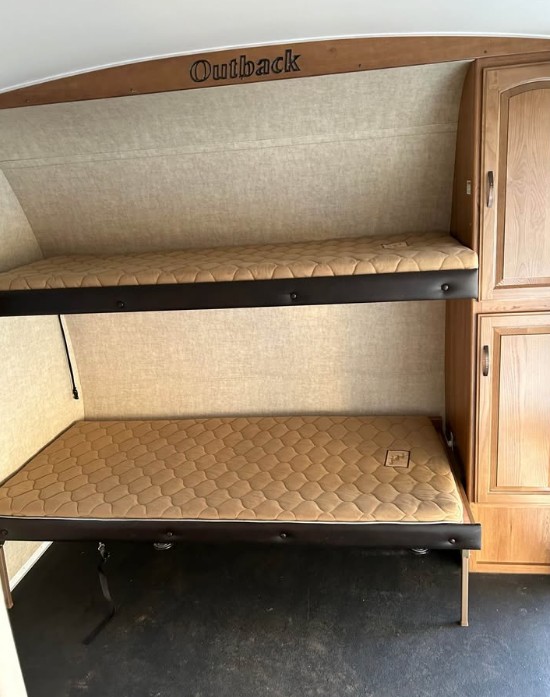 RV rental interior showcasing two stacked bunk beds with quilted tan mattresses. Outback brand visible above, next to wooden cabinets.. Keystone RV Outback Toy Hauler 2013