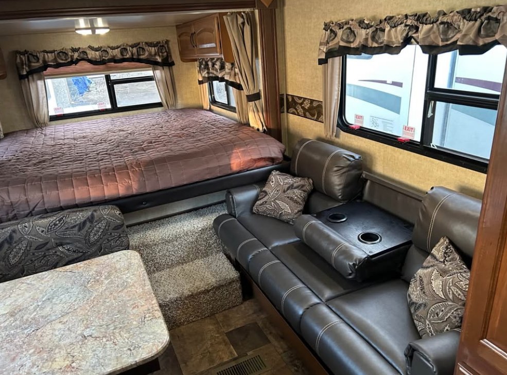 Inside this rv rental: a large bed with brown quilt, a dark sofa with cupholders, and a small table. Ready for travel comfort.. Keystone RV Outback Toy Hauler 2013