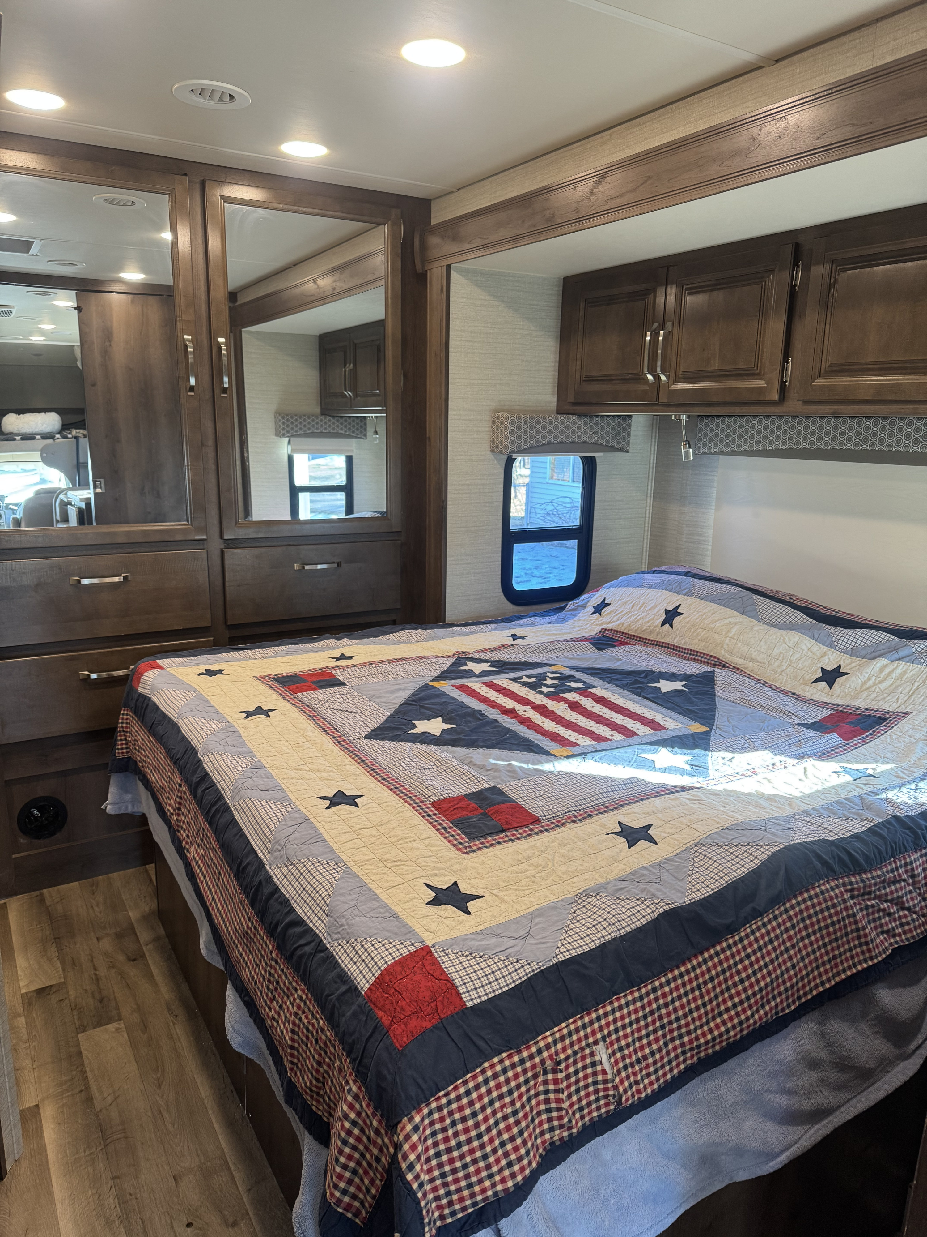 RV rental bedroom with a patriotic quilt, wood-look cabinets, mirrors, and a window, creating a cozy travel space.. Jayco Greyhawk 2020