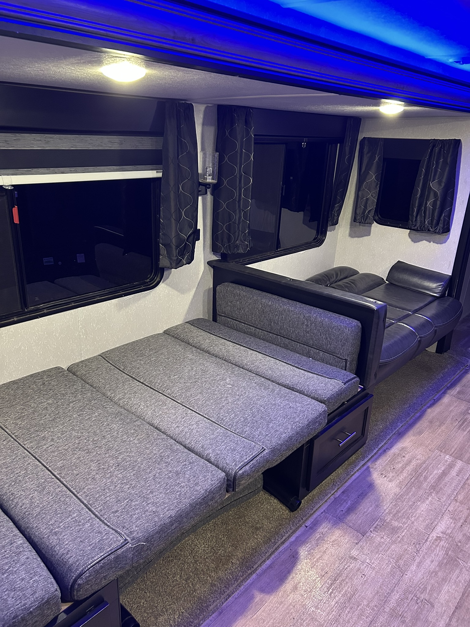 RV rental interior with a grey fold-out bed, black leather sofa, and bright blue overhead lighting, enhancing the modern feel.. Forest River Cherokee 2021