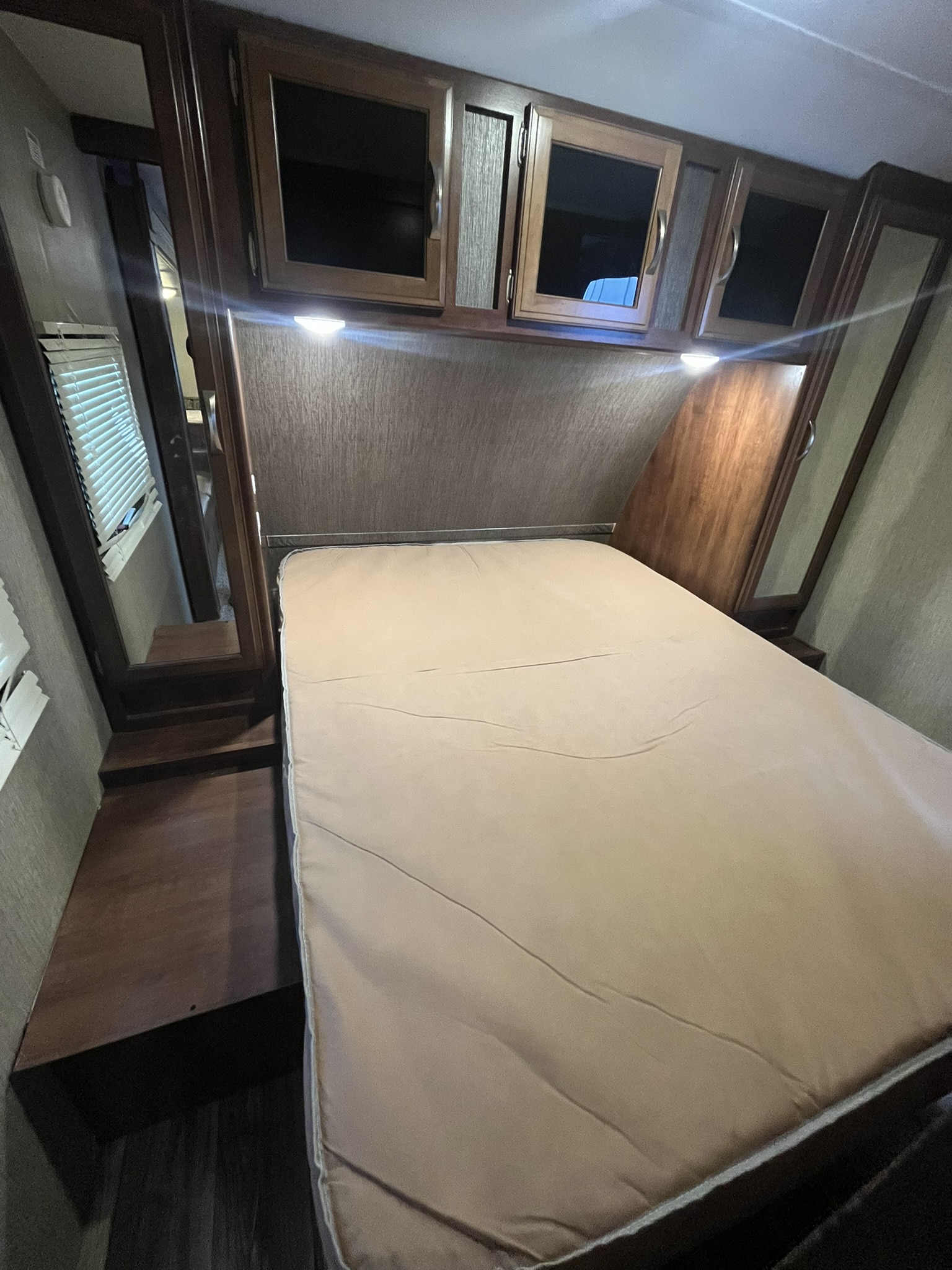 Spacious RV rental bedroom with a tan mattress, overhead wooden cabinets with lights, and integrated side storage. Mirrors reflect the interior.. Keystone RV Passport Grand Touring 2017