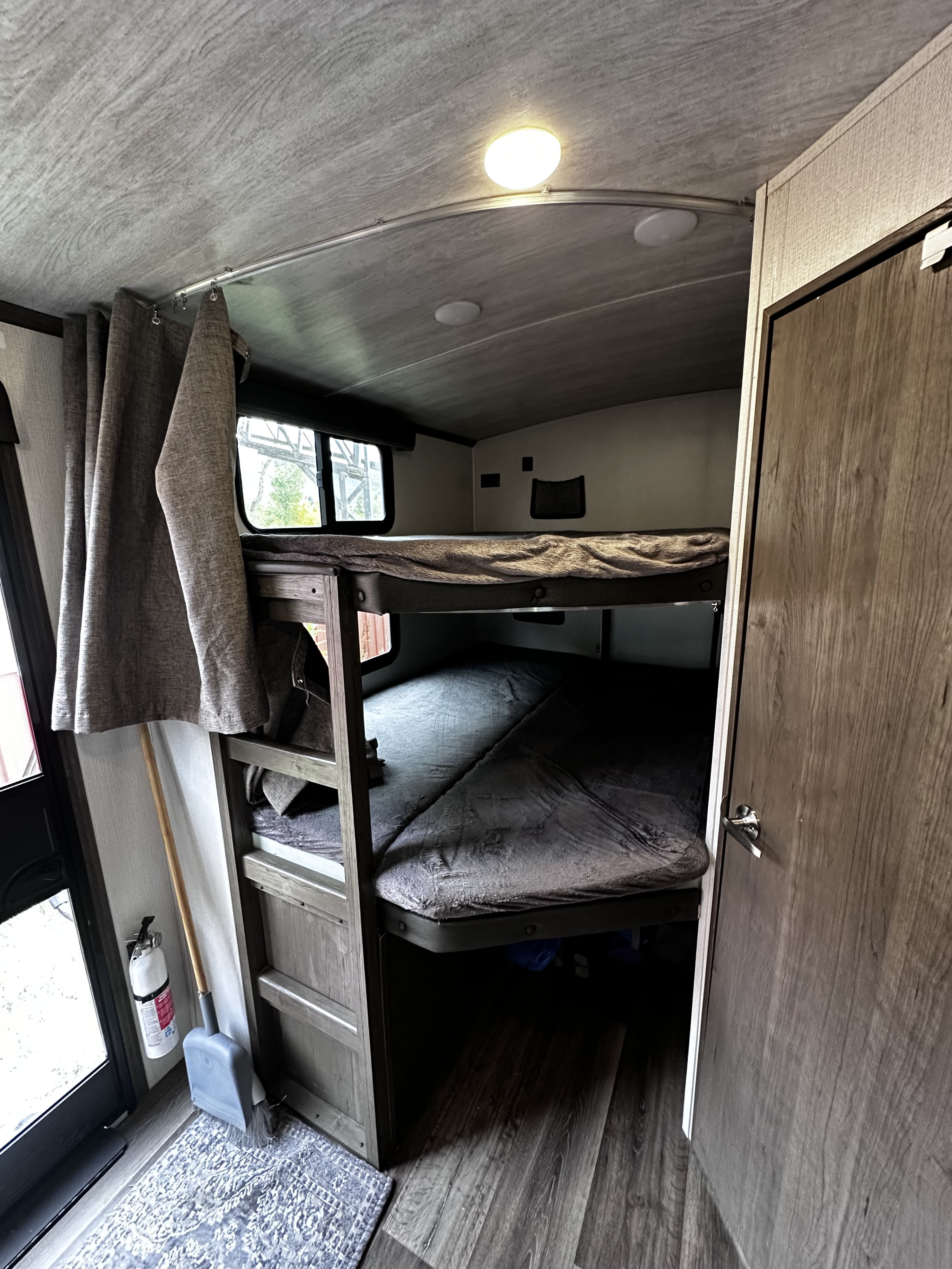 Cozy RV rental bunk beds, privacy curtains pulled aside. Features a fire extinguisher, broom, and ceiling light for comfy travel.. Keystone RV Cougar 2023