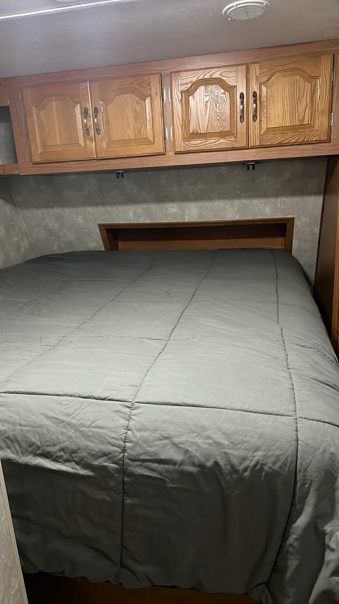 RV rental bedroom with a grey comforter on the bed, a dark wooden headboard, and light wood overhead cabinets.. Coachmen Chaparral 2004
