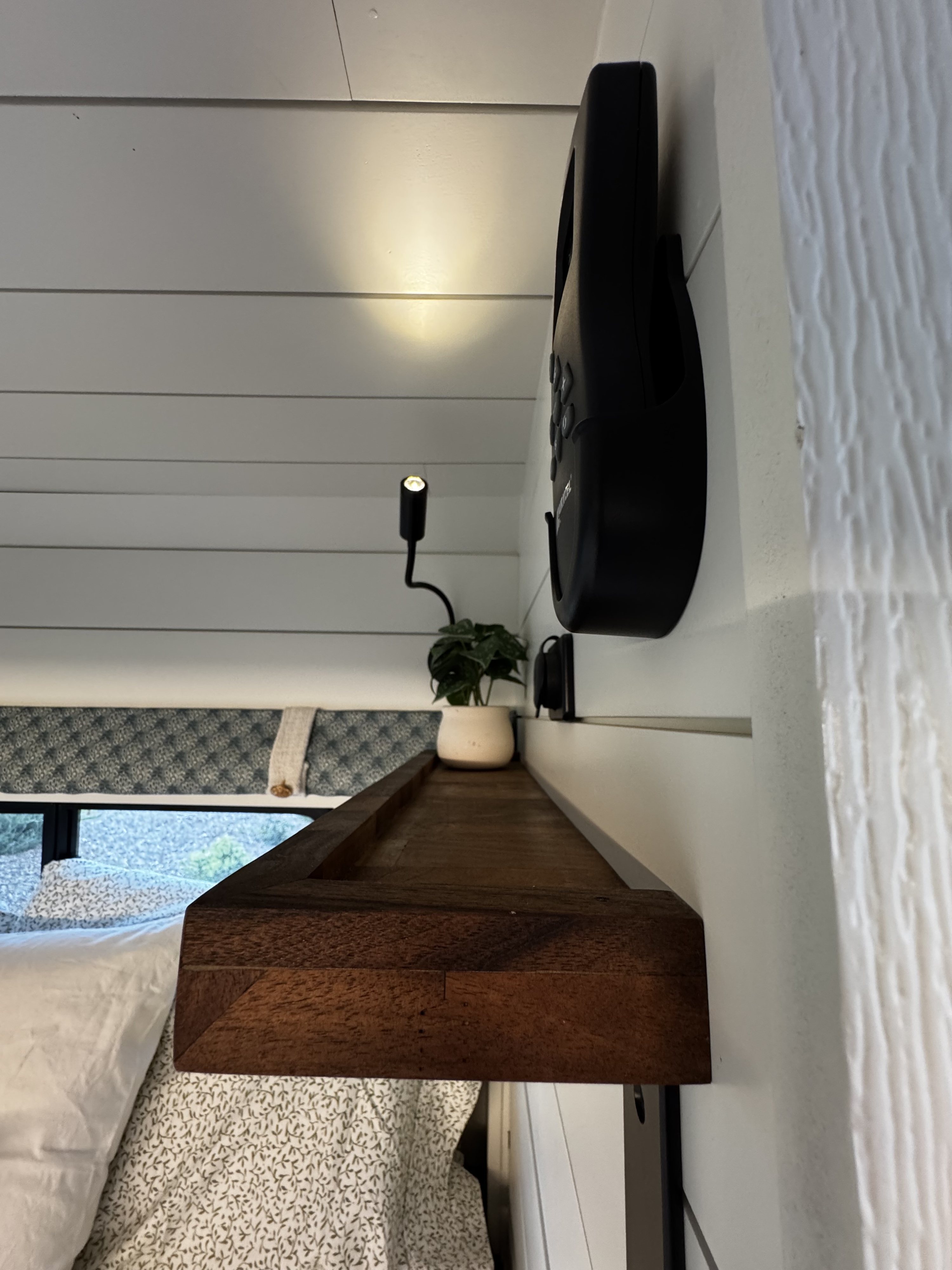 Cozy RV rental interior: wooden shelf, gooseneck light, potted plant, and a comfortable bed. Modern design.. Dodge Pro Master 2023