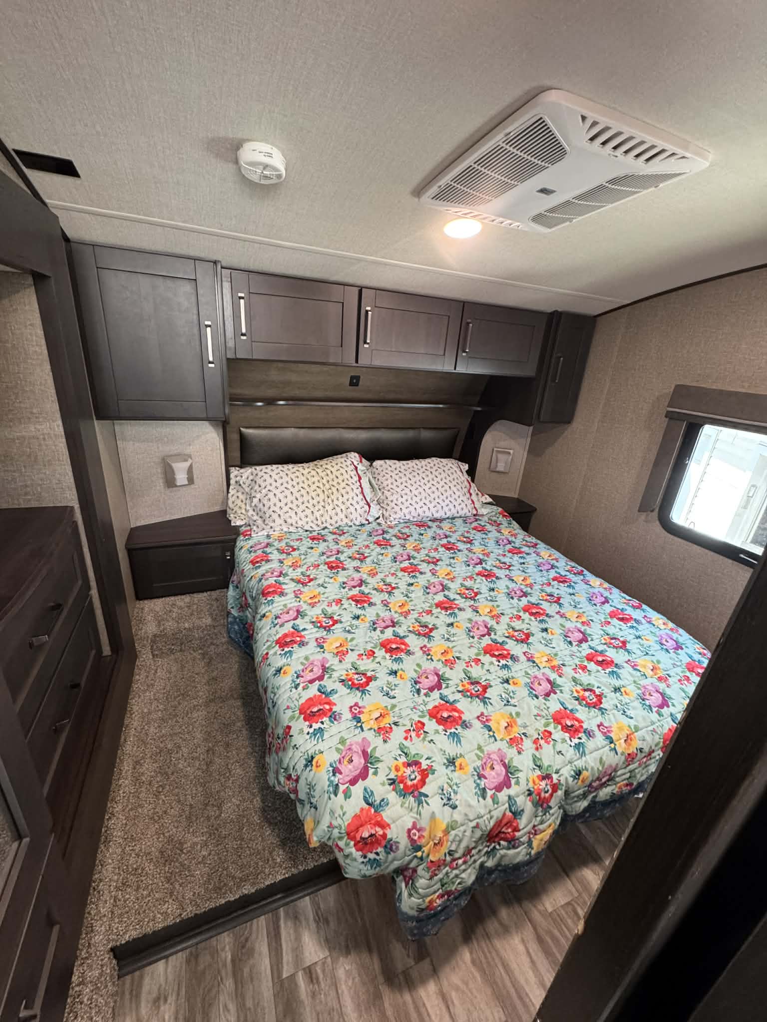Cozy RV rental bedroom featuring a vibrant floral bedspread, overhead cabinets, window, and ceiling AC unit for comfort.. Grand Design Reflection 2022