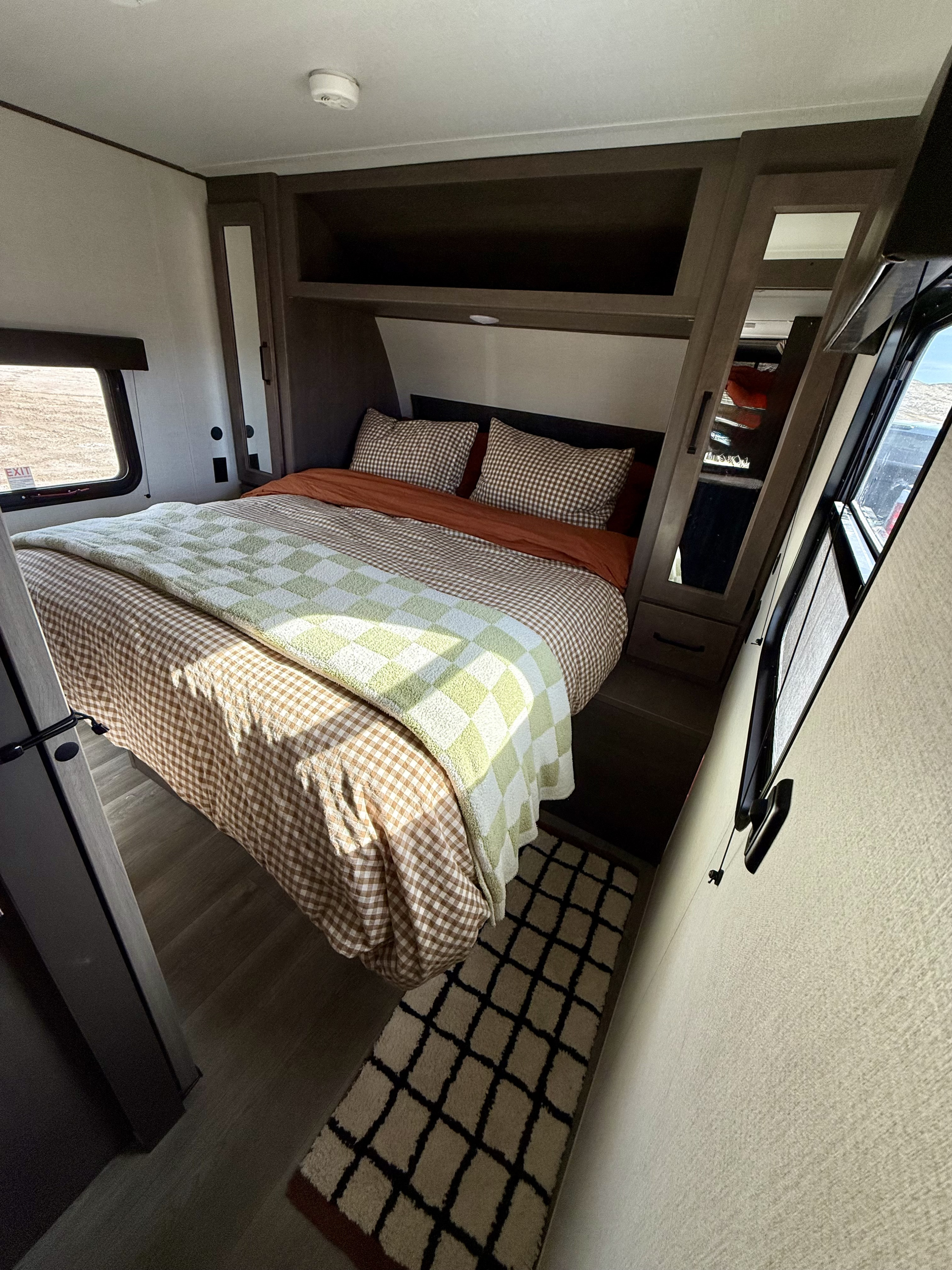 Inside a bright RV rental bedroom with a brown gingham and green checkered bed, built-in storage, and sunlit windows.. Grand Design Transcend Xplor 2024