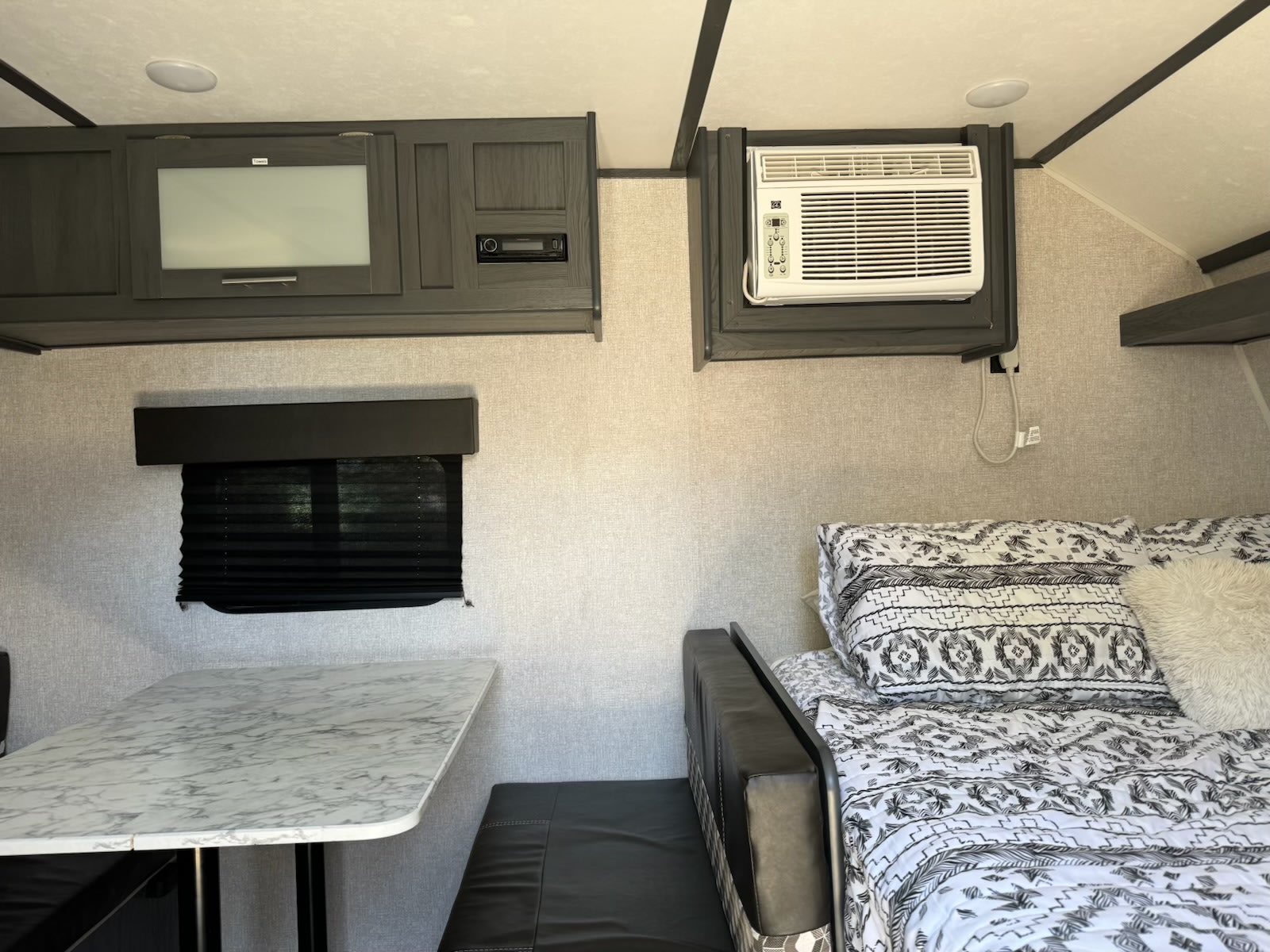 Interior of an RV rental: cozy bed with patterned bedding, marble-look table, window, and wall-mounted AC unit and TV/media center.. Aspen Trail Aspen Trail Trailer 2022