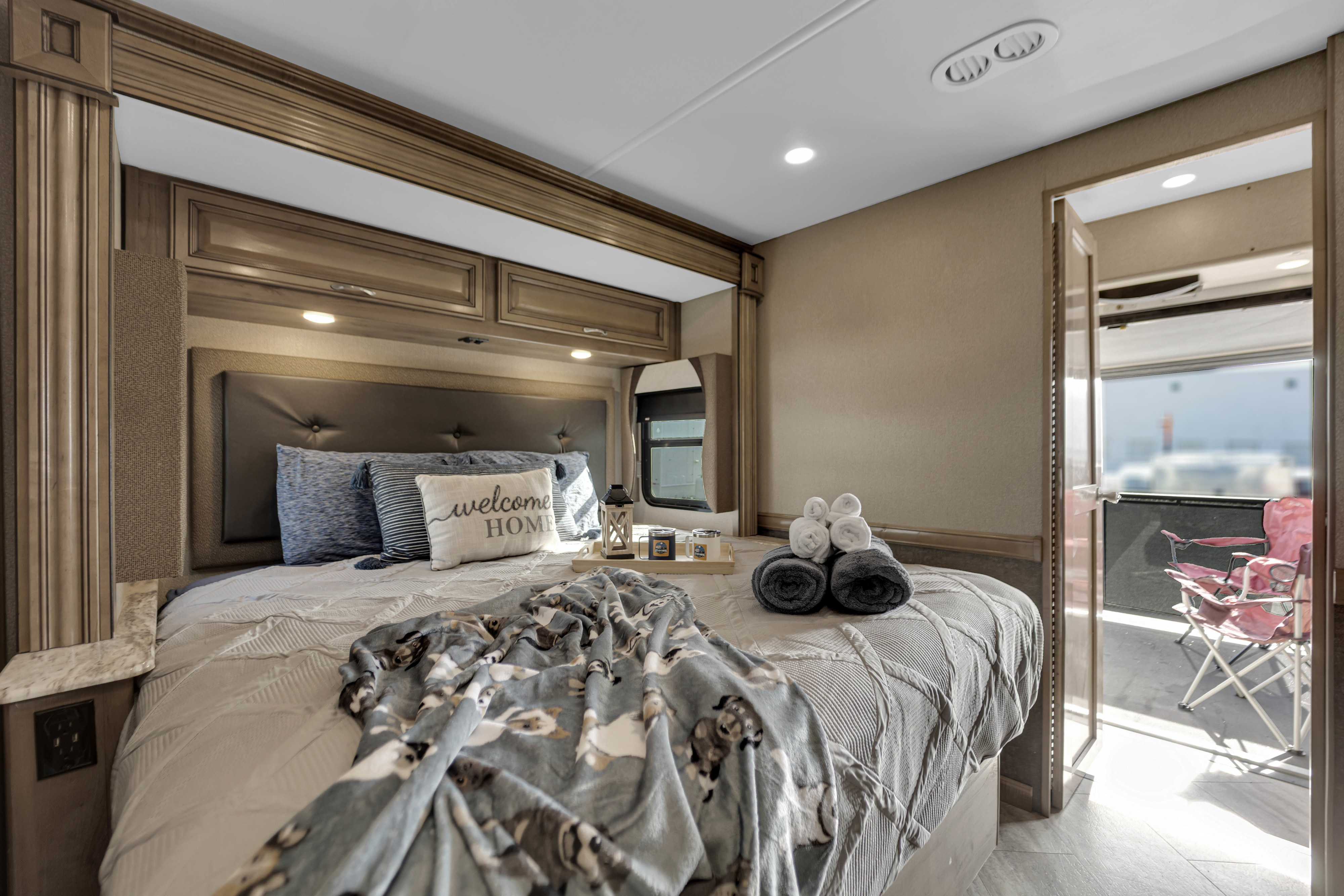 Cozy RV rental bedroom with a large bed, "welcome home" pillow, and blanket, offering a glimpse of an outdoor patio with pink chairs.. Fleetwood Bounder 2019