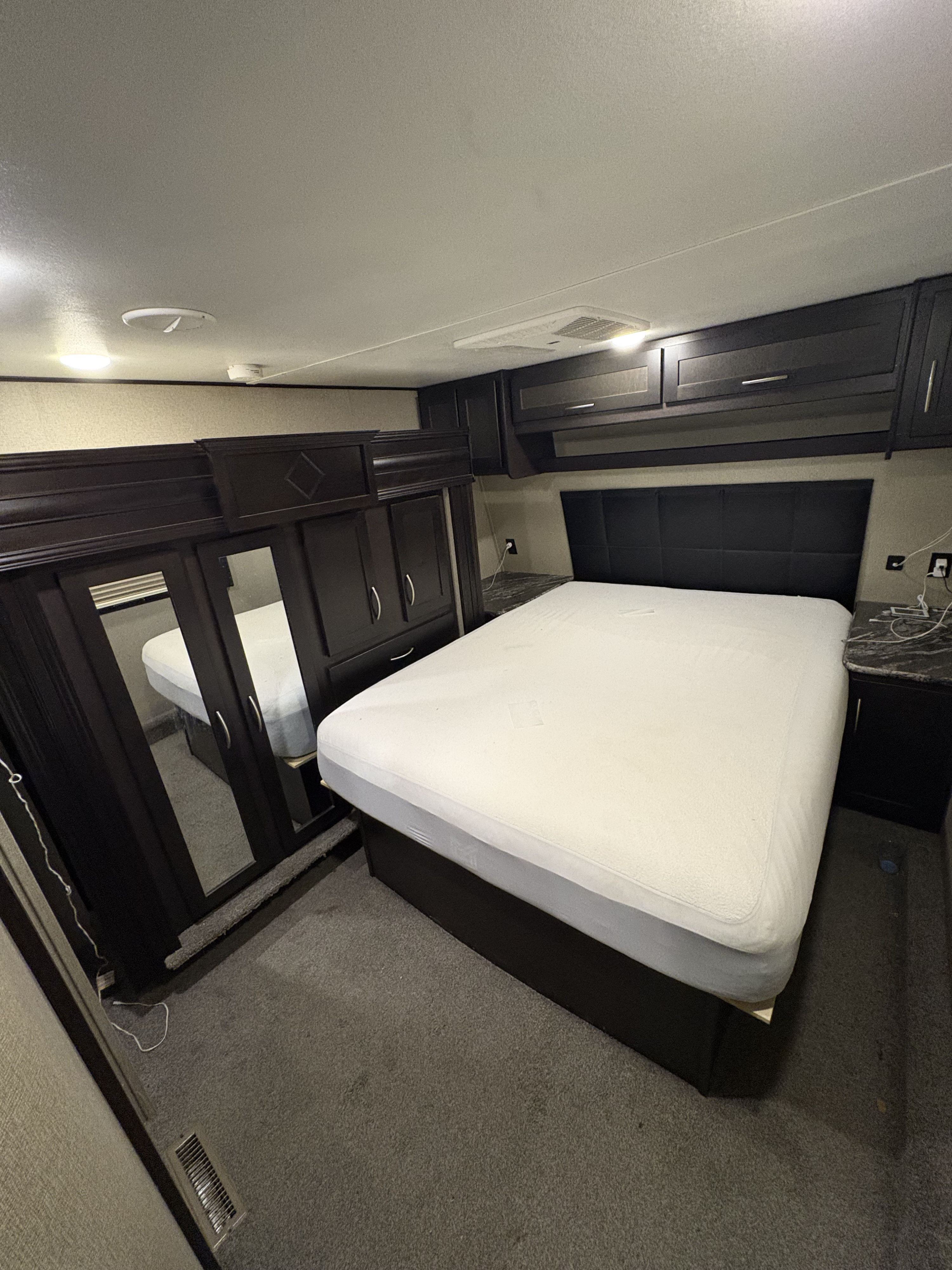 RV rental bedroom interior: queen bed with dark headboard, dark wood wardrobes with mirrors, overhead cabinets, and carpeted floor.. Keystone RV Carbon Toy Hauler 2018
