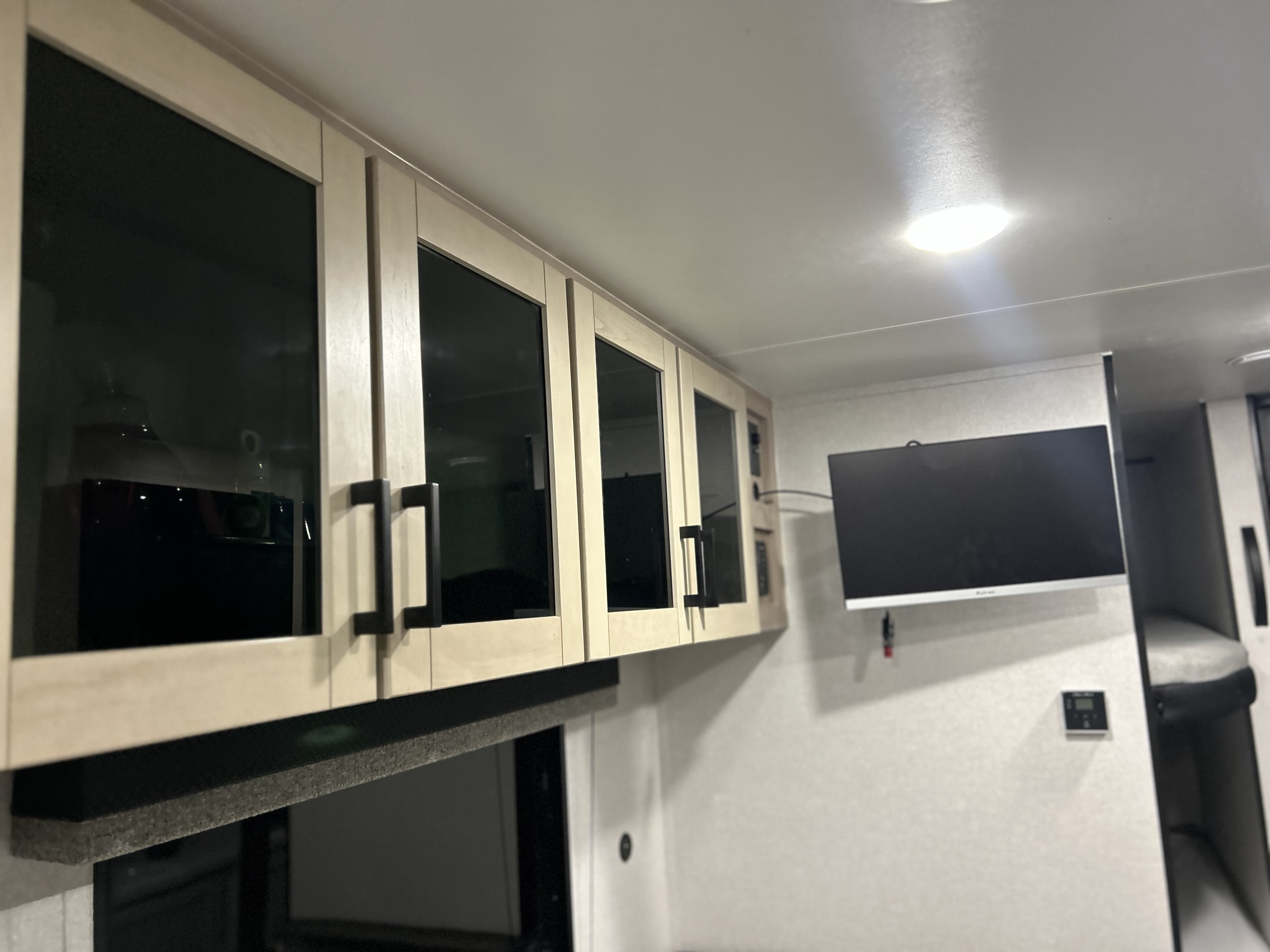 Inside an **rv rental**: Light wood cabinets with dark glass doors and black handles, a wall-mounted TV, and bright ceiling lights.. Jayco Open Range 2026
