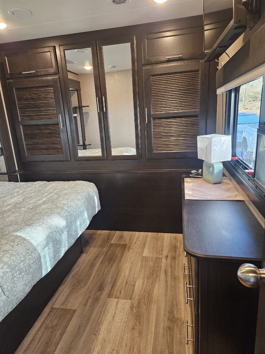 Inside an RV rental, this bedroom features dark wood cabinetry, mirrored closets, a bed, a dresser with a lamp, and wood flooring.. Jayco Eagle 2021
