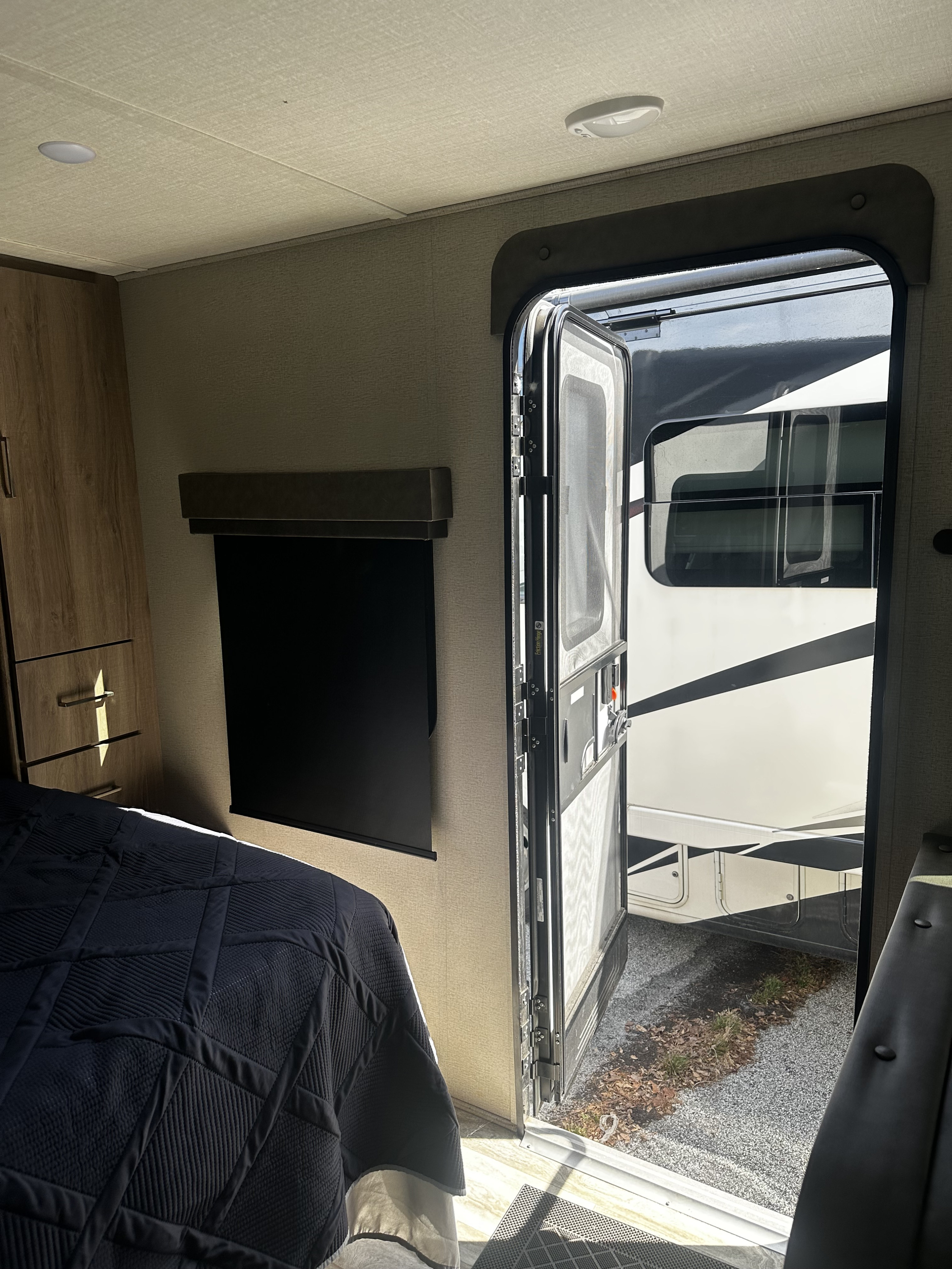 Interior view of an RV rental with a dark blue bed, wooden cabinet, and an open door revealing the camper's exterior and gravel ground.. Grand Design Imagine XLS 2021