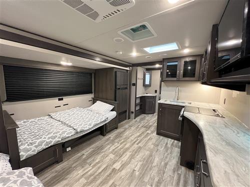 RV rental interior with a cozy bed, dark blinds, and a well-equipped kitchen featuring a counter, sink, and TV.. Grand Design Transcend Xplor 265BH 2021