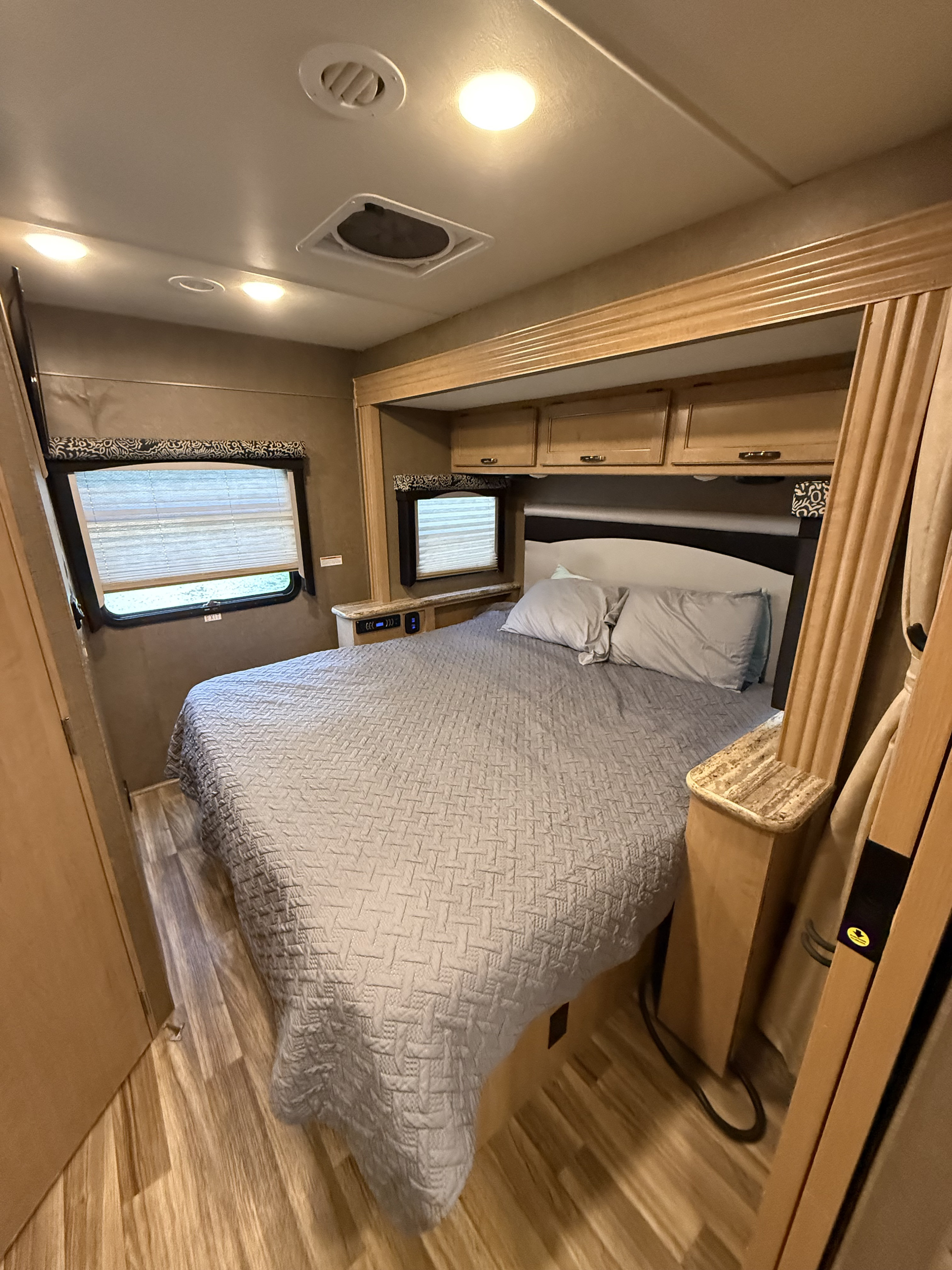 Comfortable RV rental bedroom featuring a quilted bed, windows with blinds, and overhead storage, perfect for travel.. Thor A.C.E. 2018
