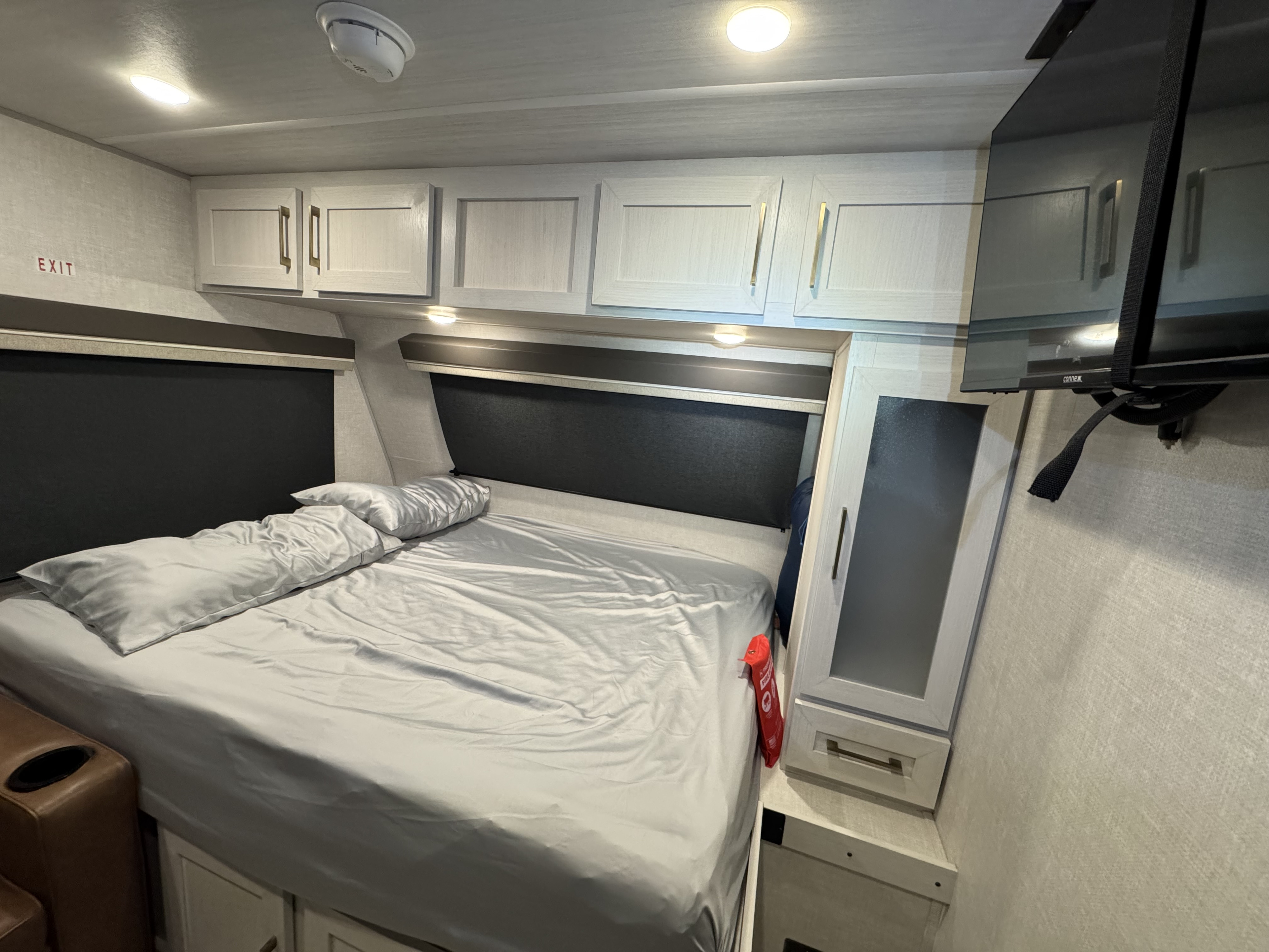 Cozy RV rental bedroom with a light gray bed, overhead cabinets, wall-mounted TV, and an exit sign visible on the left.. Forest River No Boundaries 18.0 with beastmode 2025