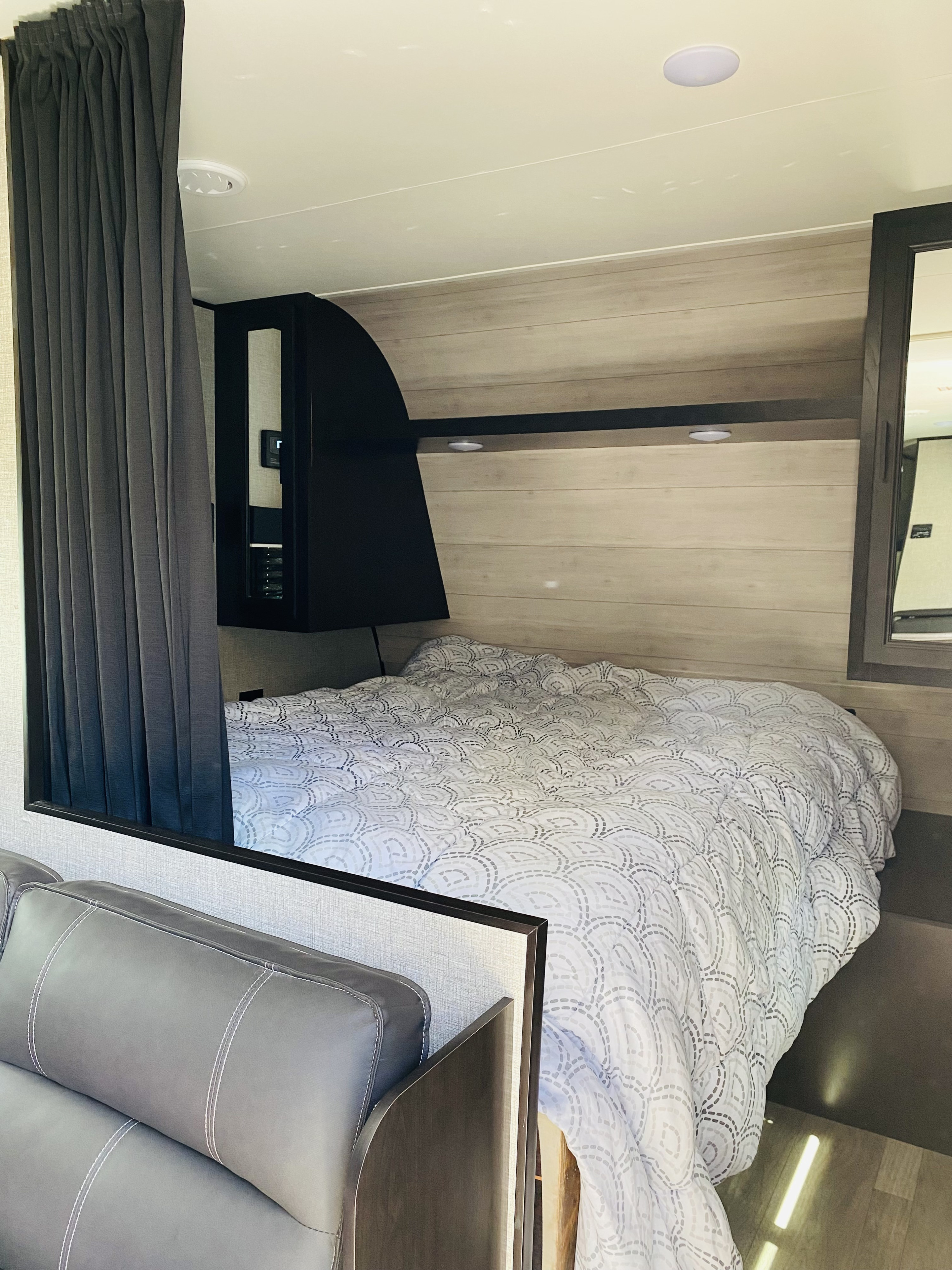 Cozy RV rental bedroom with a patterned bed, dark privacy curtains, and wall storage. A grey sofa armrest is visible.. Jayco Jay Flight SLX Rocky Mountain Edition 2022