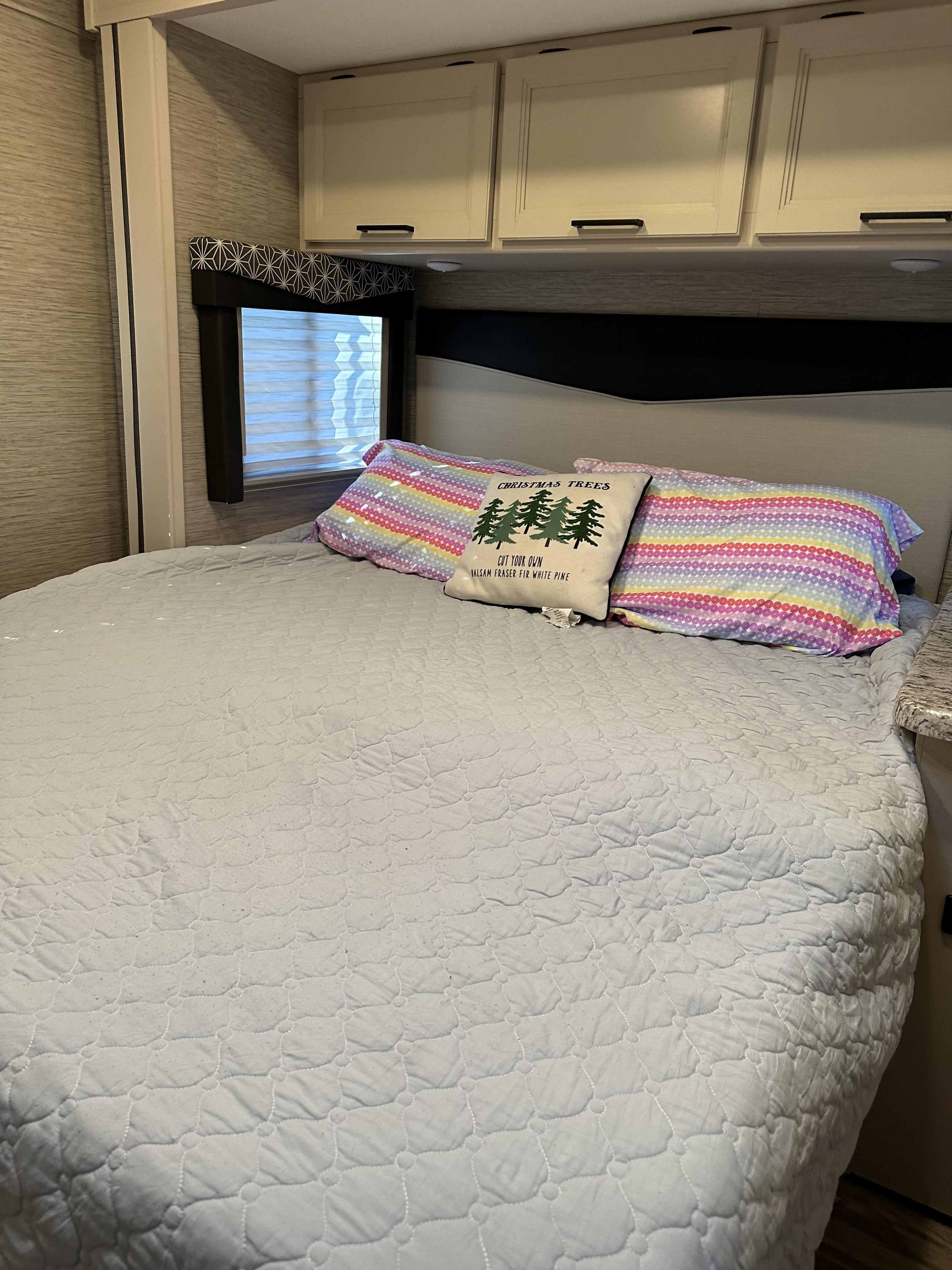 RV rental bedroom: a soft bed with colorful pillows, a Christmas tree pillow, window, and overhead cabinets.. Ford E350 2023