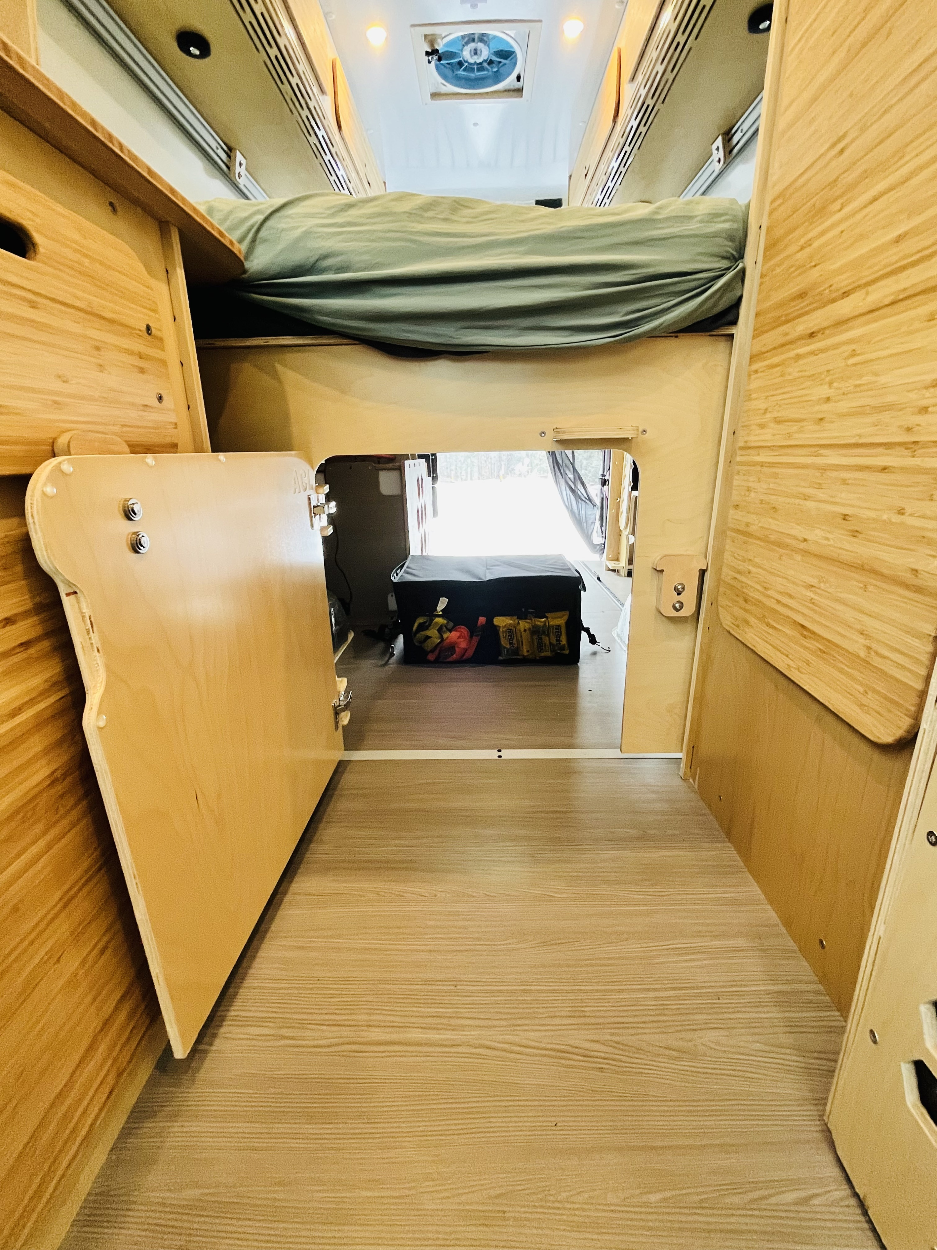 Cozy RV rental interior featuring an overhead bed, under-bed storage with a cooler, and light wood paneling.. Mercedes Sprinter 2015