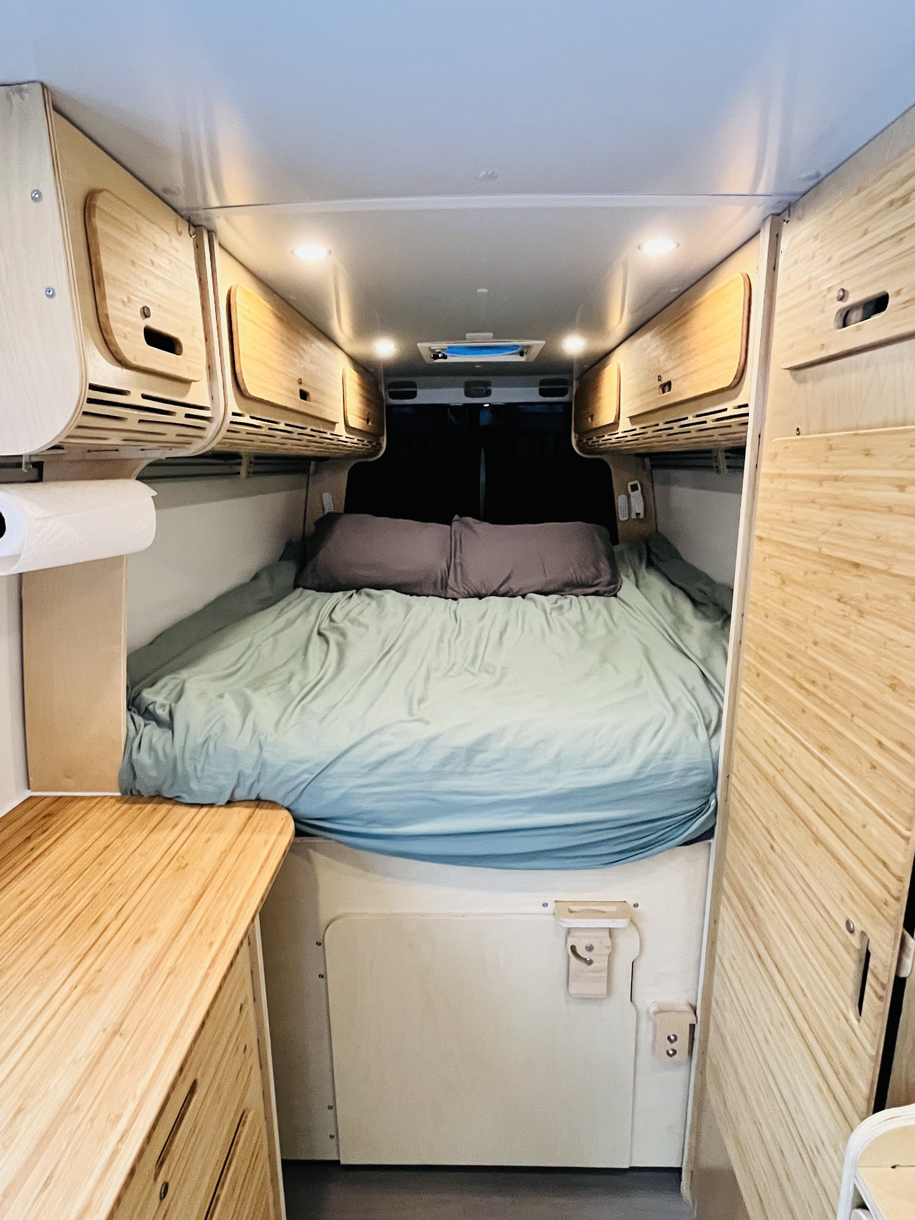 Cozy RV rental interior featuring a comfortable bed with green bedding, overhead wooden cabinets, and bright ceiling lights.. Mercedes Sprinter 2015