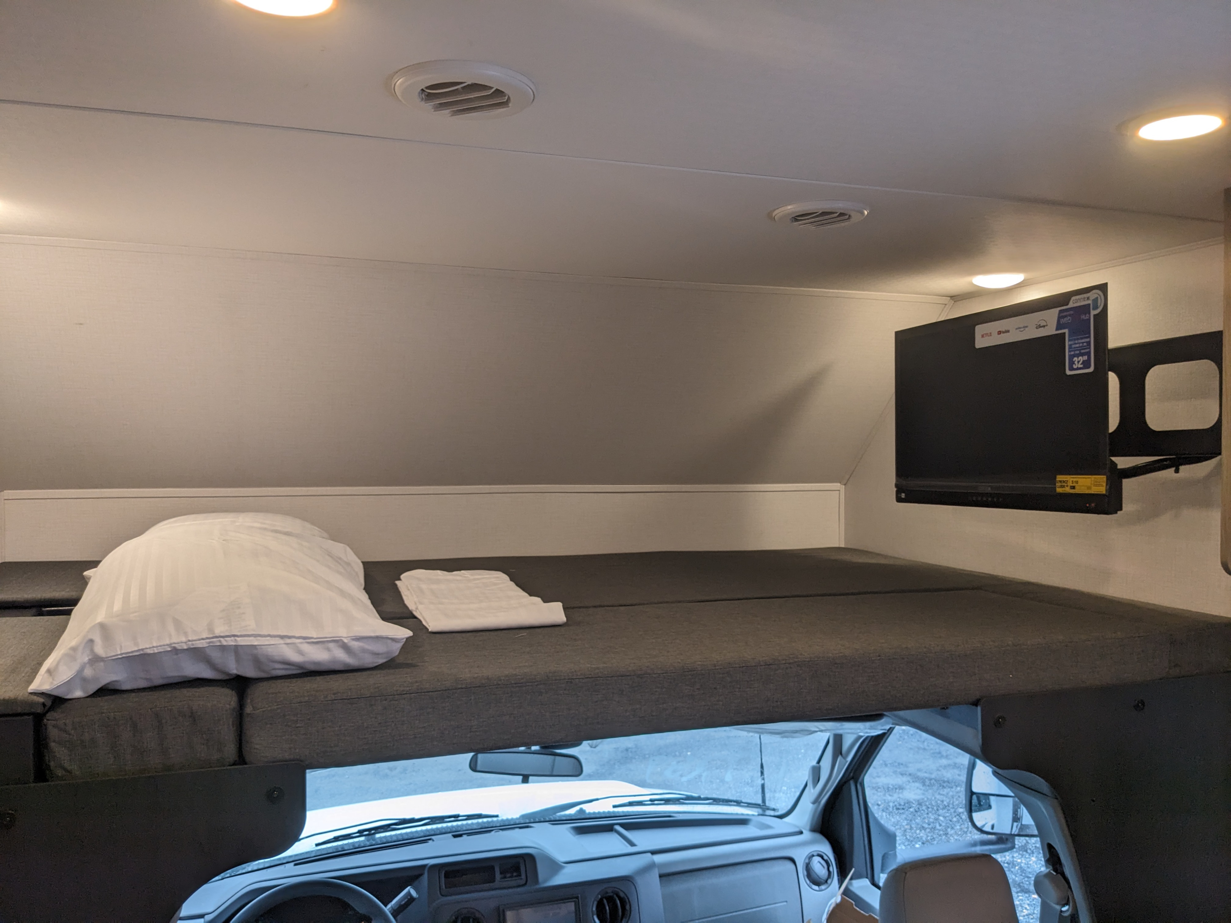 Inside a cozy RV rental: a cab-over bunk bed with pillow & linens, TV, and the vehicle's dashboard visible below.. Forest River Forester 2027