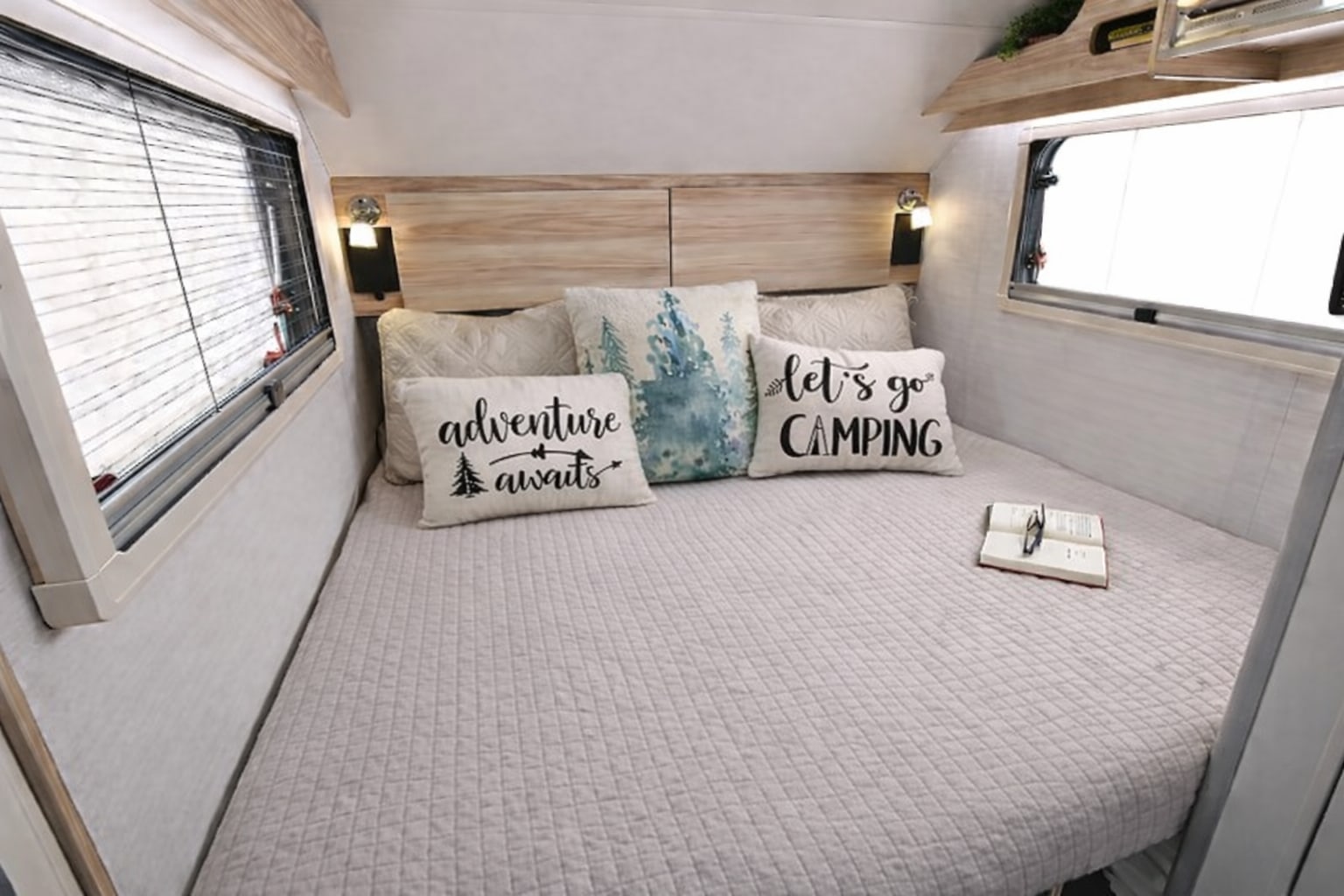 Inside a comfortable RV rental: a neatly made bed with camping-themed pillows, large windows, and an open book. Ideal for trips.. nuCamp T@B 2024